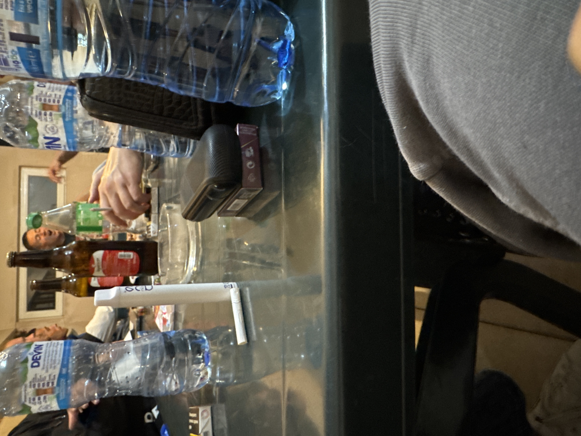 A cluttered tabletop scene in Topolovets, Bulgaria, reveals several individuals around a dark, possibly green, table. Two individuals are visible, while others are partially shown. One person wears a gray sweater and sits at the table. Another person is shown lower down in the frame, wearing a black top with a white logo.
The table is covered with various items including multiple plastic water bottles (both large and small), glass bottles, a white vaping device, a wallet, and a pack of cigarettes. Empty glasses and a clear pitcher are also present.
The location is Topolovets, Bulgaria, suggested by the "Devin" water bottle, a brand associated with Bulgaria. The atmosphere suggests a casual gathering or social event. The time of day is unclear, given the indoor lighting, although there is a window in the background.
