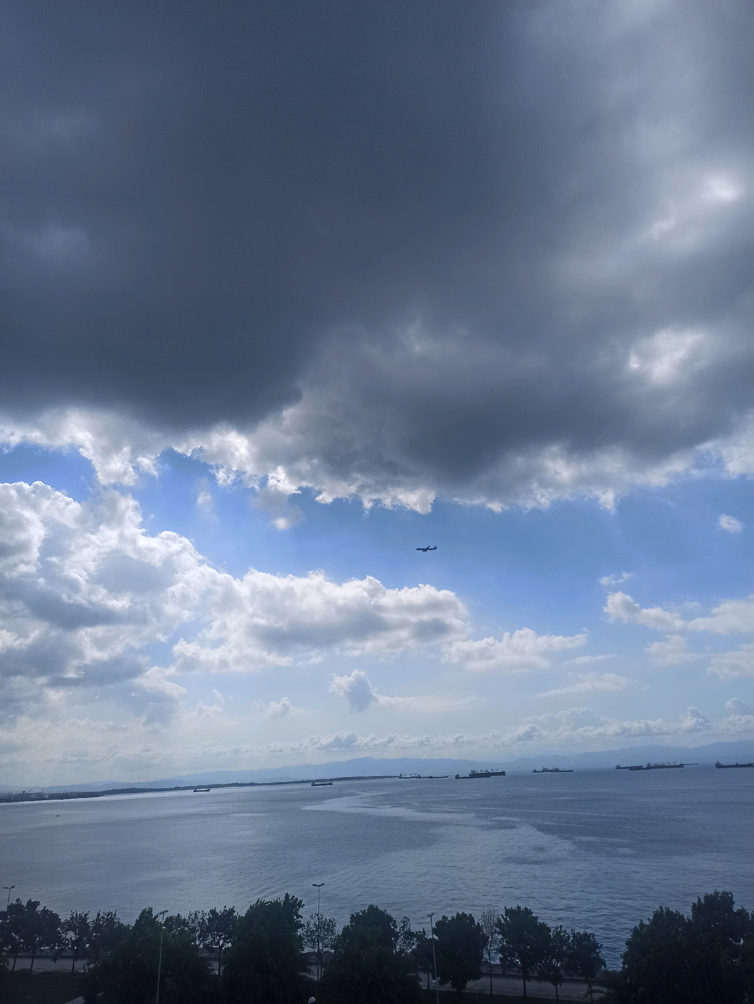 An airplane flies across a wide expanse of sky above a body of water. The sky is filled with a mix of dark, ominous storm clouds and patches of bright blue sky with fluffy white clouds. In the distance, a line of cargo ships is visible on the water, and a distant coastline with mountains can be seen. The foreground of the image shows a row of dark trees along a waterfront, with streetlights interspersed among them. This scene is consistent with the views from Pendik, Turkey, which is situated along the Sea of Marmara and is known for its maritime traffic.