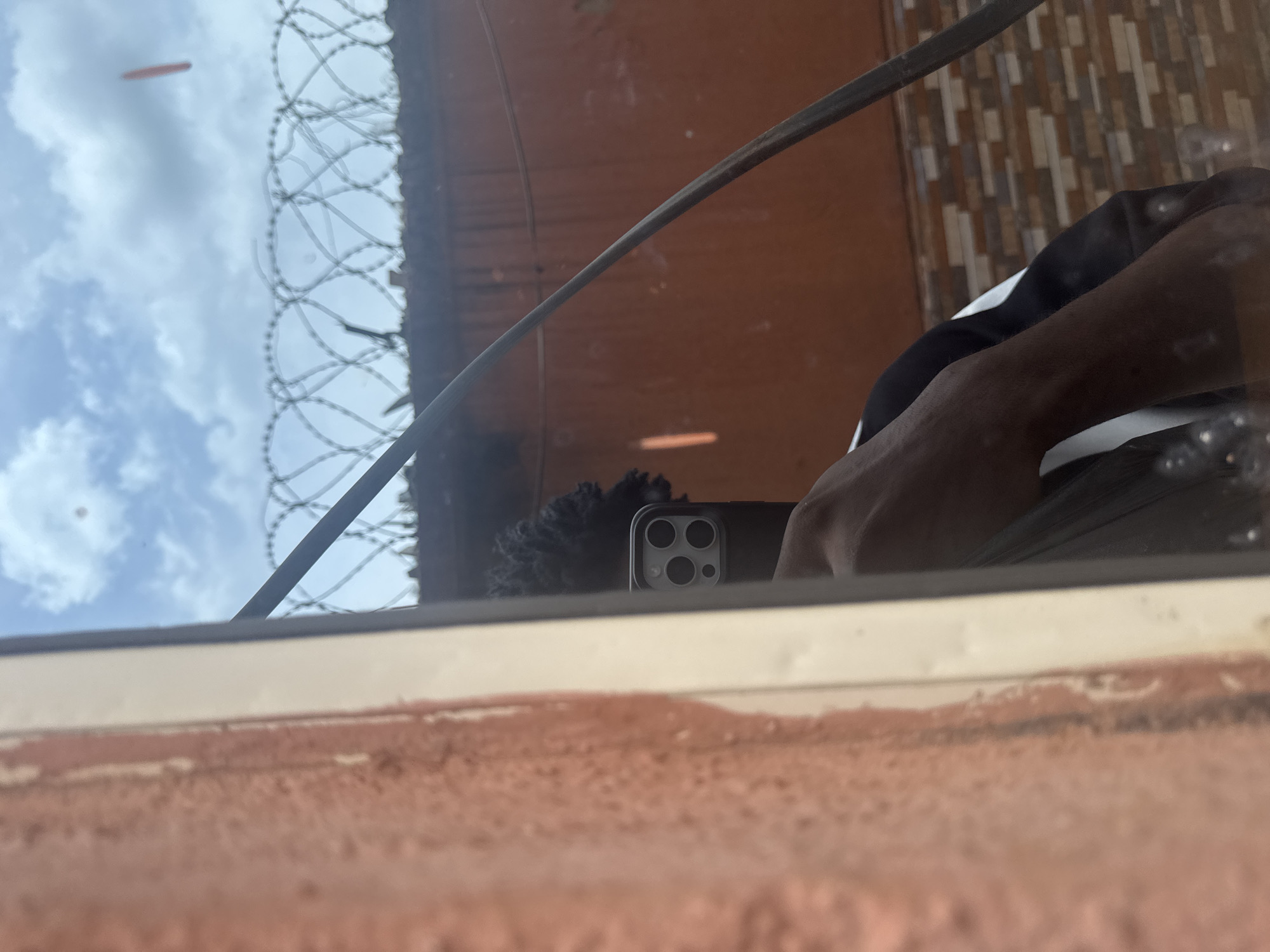 The photograph, taken from what appears to be inside a building in Ihiagwa, Nigeria, shows a reflection in a window. The reflection shows an individual holding a smartphone, presumably taking the photo. The subject's skin tone appears to be dark. The smartphone is black with a triple-lens camera. The background as seen through the window consists of the sky with clouds and an orange wall with barbed wire on top. A cable runs diagonally across the scene. The window's frame is partially visible at the bottom of the image.