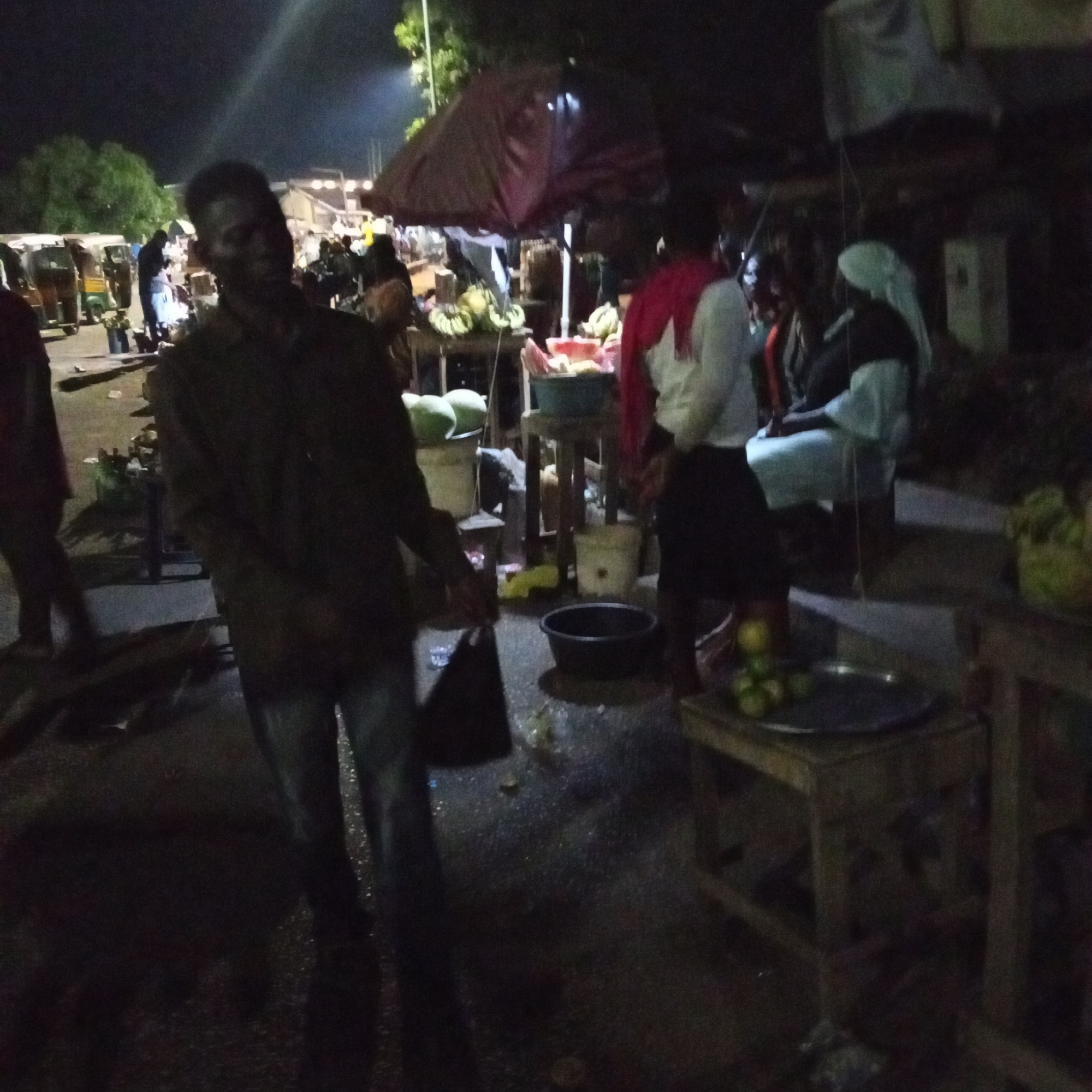 The image depicts a street scene at night in what appears to be a market area in Unknown, Unknown. Several people are present, including a man in the foreground wearing a dark jacket and jeans. He is looking at the camera. In the background, vendors are selling goods under a canopy, including what seems to be produce like bananas, watermelons, and fruits. There are some seated individuals wearing white head coverings. Lighting is poor, with areas in shadow. Other notable objects include various wooden tables, a black bucket, and an assortment of items displayed for sale.