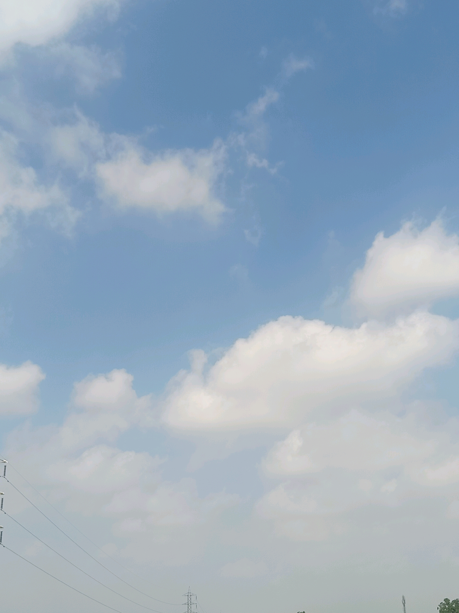 The image captures a wide-angle view of a bright, overcast sky with scattered white clouds. The lower portion of the image reveals the tops of utility poles and power lines, characteristic of infrastructure found in a rural or semi-urban environment. The overall scene suggests a typical daytime atmosphere in Kaluwala, India, with a focus on the sky and the presence of man-made structures.