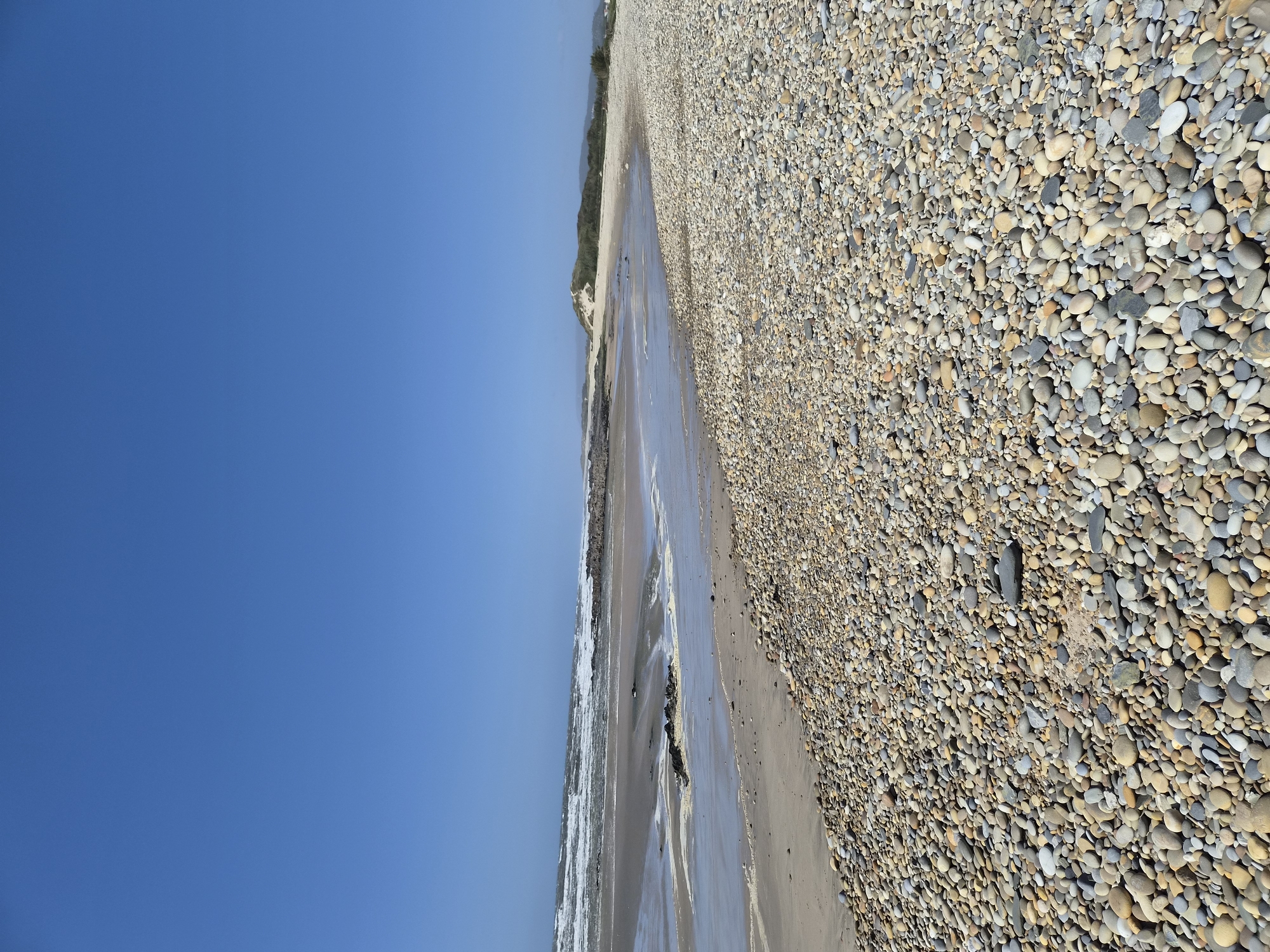 The image depicts a beach scene in Esposende, Portugal. A large expanse of sky occupies the left portion of the image, transitioning to a coastline. The beach itself has a wide stretch of sand, partially covered with water, indicating the ebb of the tide. A substantial area to the right is composed of small, smooth pebbles, densely packed together. Further in the distance, vegetation is visible along the coast.