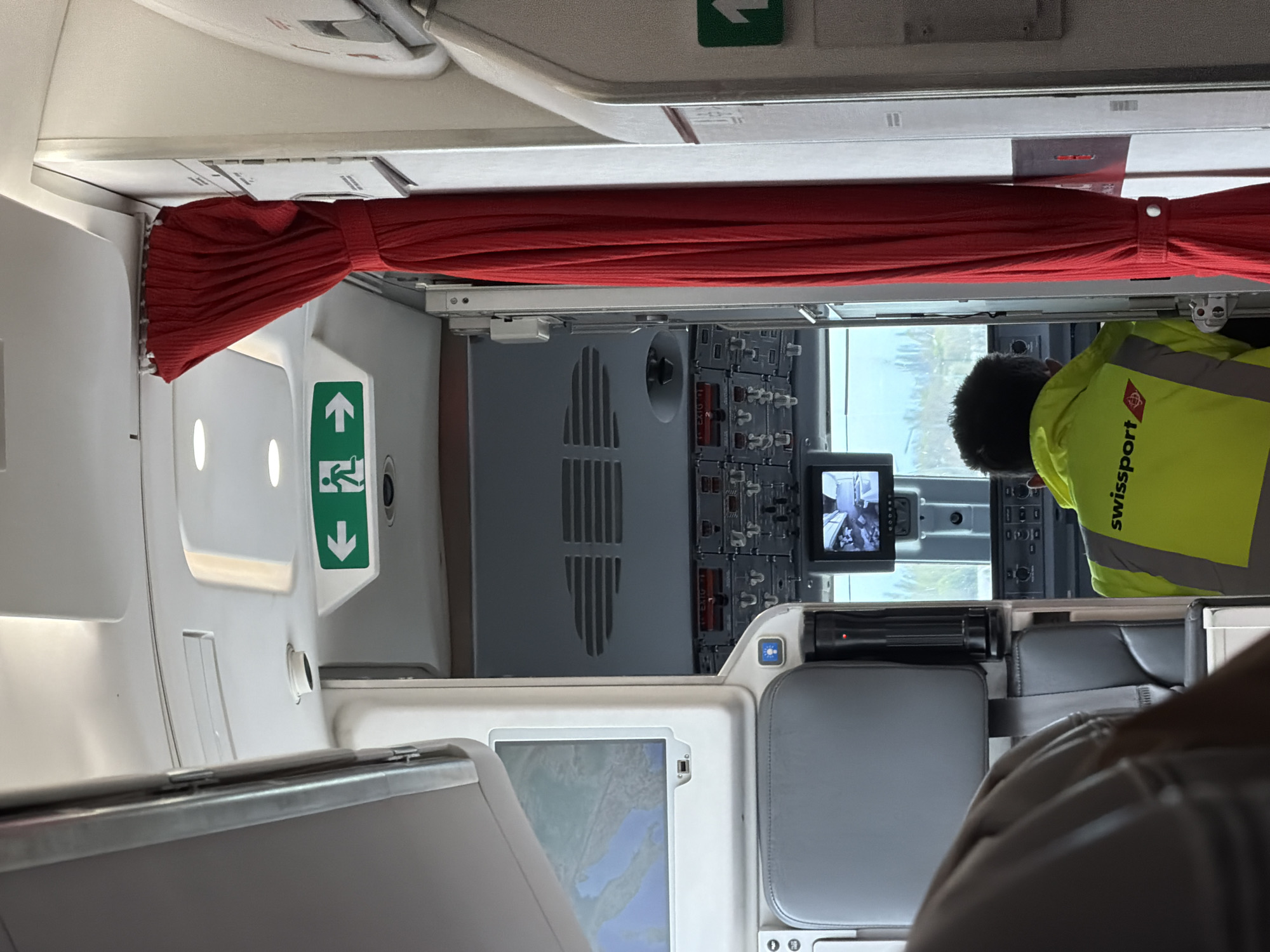 The image shows the interior of an airplane cockpit, possibly during pre-flight checks or maintenance. The view is from behind the cockpit seats, looking towards the front. A red curtain separates the cockpit from the passenger cabin. The instrument panel with various switches, gauges, and a small display screen is visible. A person wearing a bright yellow "swissport" safety vest is present in the cockpit. A seat and screen are visible in the foreground. The location cannot be determined from the image, and there is no evidence of Sofia, Bulgaria.