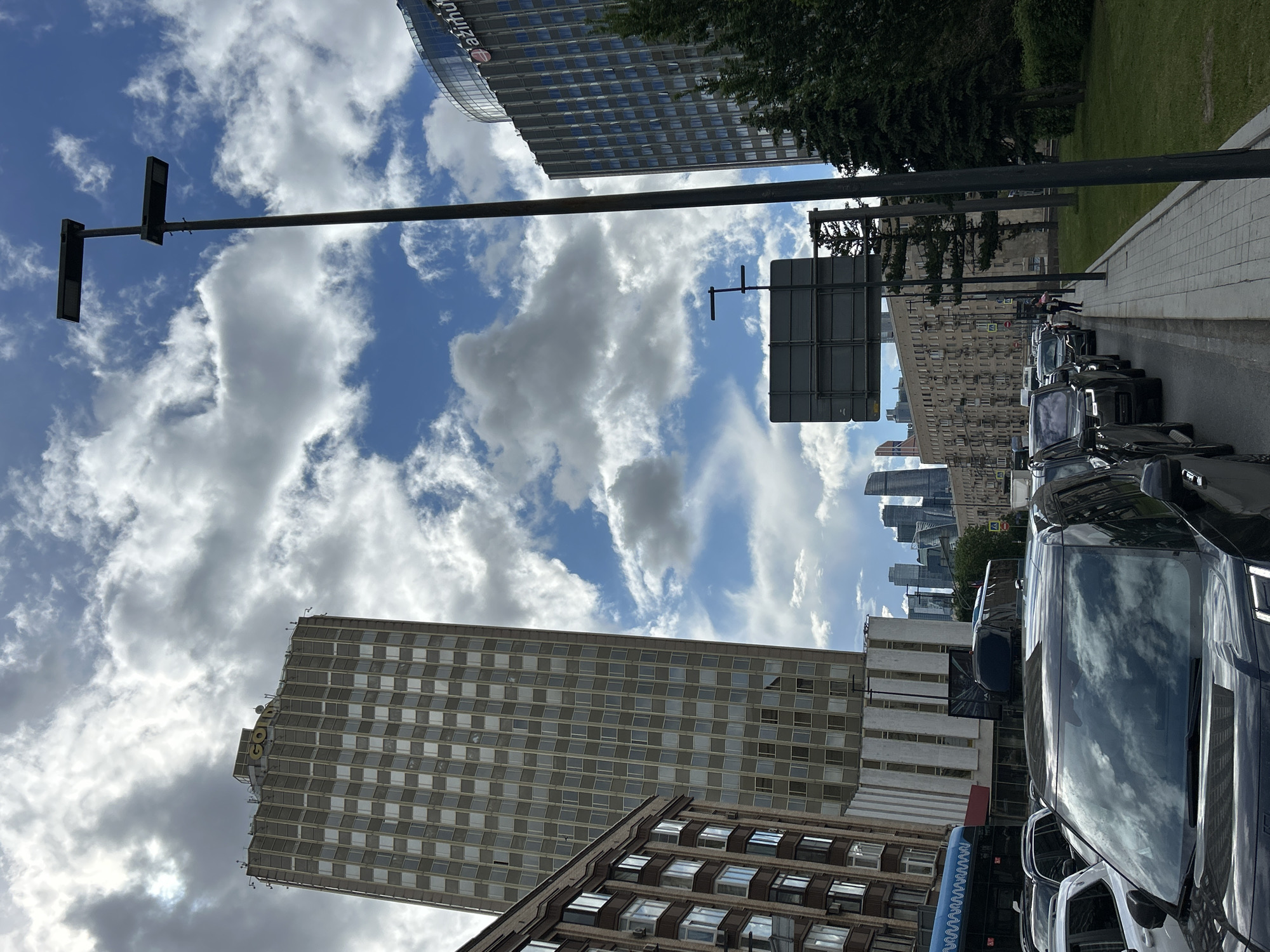 The image shows a city street with a row of parked cars on the right. The sky is visible, filled with blue patches and white, puffy clouds. Several tall buildings are visible, including one with a sign that reads "Gazprom". There is also a building that may read "GOLDEN RING" on the top left. Traffic lights and street signs are mounted on poles along the street. In the background, a cluster of modern skyscrapers is visible. People can be seen walking on the sidewalk in the distance. The exact city and location are not discernable from the image.