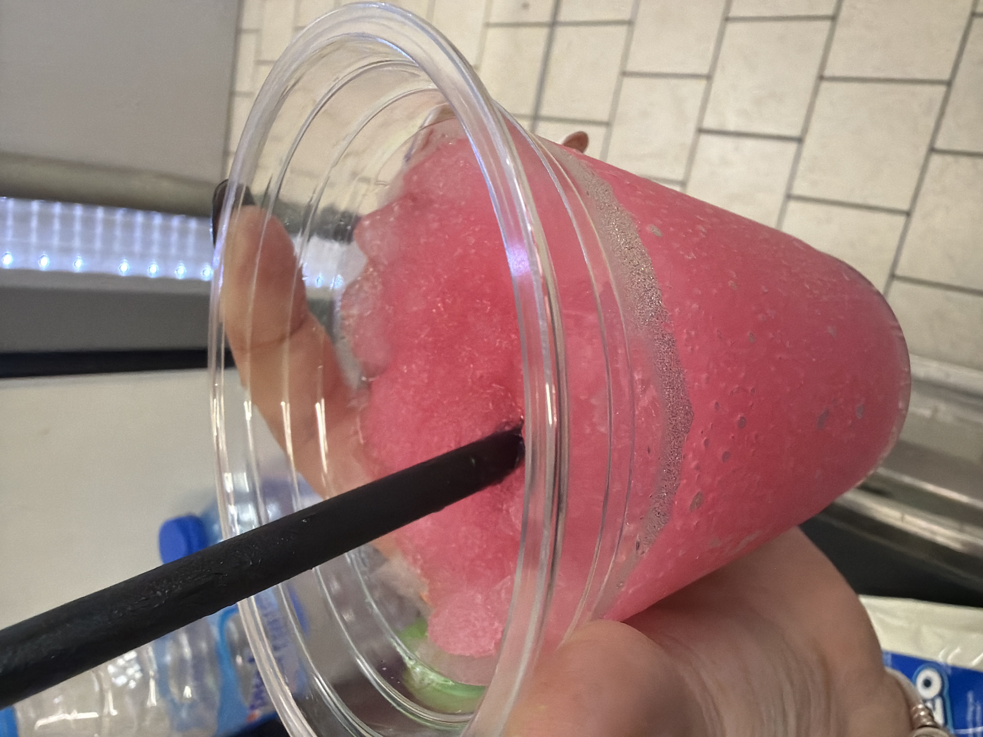 A close-up, tilted shot captures a clear plastic cup filled with a vibrant pink, slushy-like beverage, featuring visible ice crystals and condensation on its exterior. A black straw is inserted into the frozen drink. A hand, with light skin tone and wearing a ring on one finger, holds the cup.
The background elements suggest an indoor setting, potentially a casual commercial space or a home in Plovdiv, Bulgaria. To the left, a strip of bright LED lights provides illumination. Behind and above the cup, a wall or counter surface is visible, covered in light-colored, possibly beige or white, square tiles. Further in the background, parts of plastic water bottles with blue caps are discernible. In the bottom right corner, a white package is partially visible, showing the text "OREO".
The image focuses on the refreshing drink, implying the activity of enjoying or about to enjoy a cold beverage. The time of day and weather are not evident, but the nature of the drink suggests a need for refreshment. No specific emotions are discernible from the partial view of the hand.