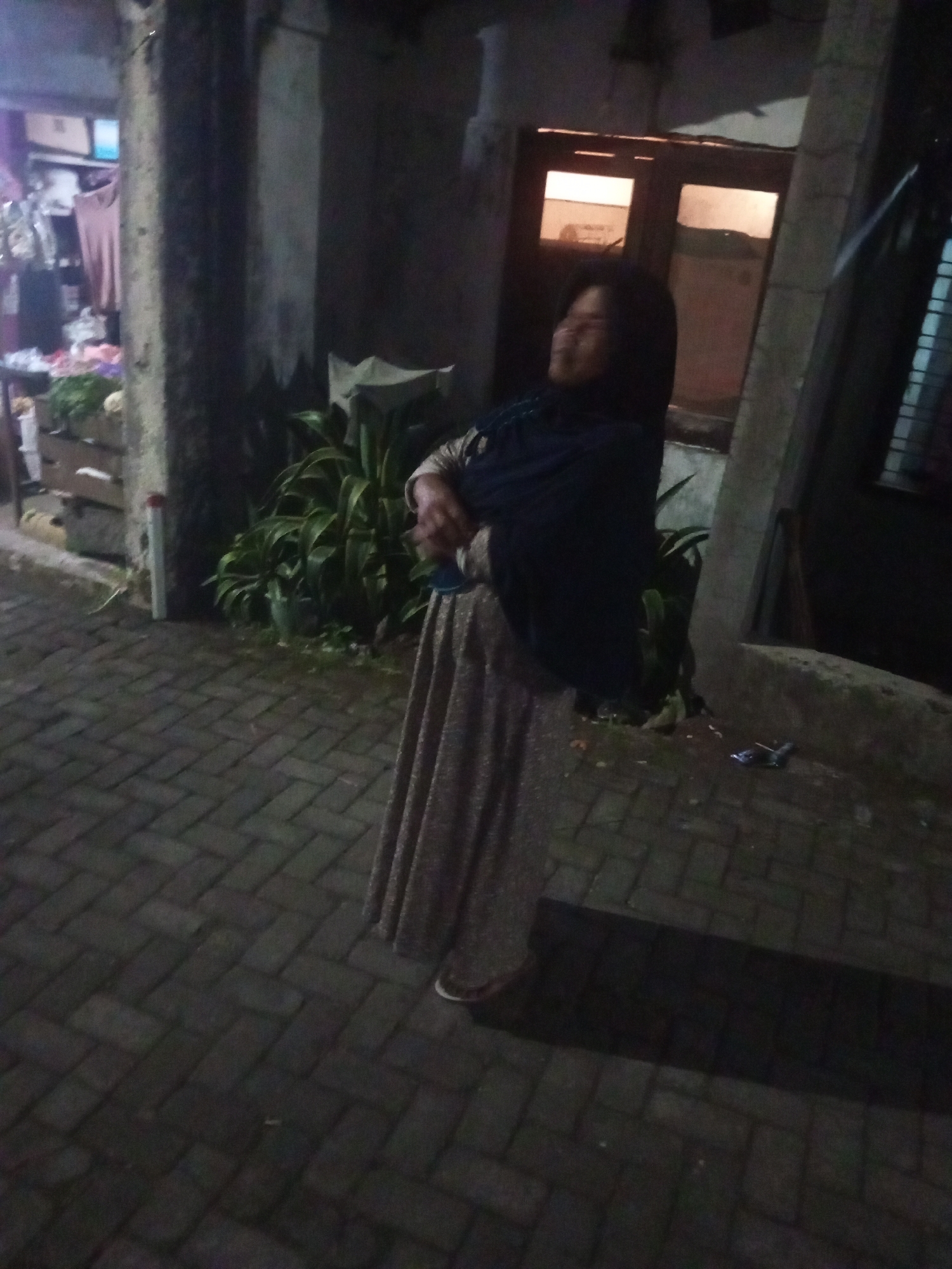 A person wearing a dark hijab and a long, patterned dress stands on a brick walkway in front of a building at night. The person is looking upwards and to the left with their eyes closed. To the left of the person is a pillar and a display of goods, possibly in a shop. Behind the person, a window with internal lighting is visible in the building. Green foliage is also present near the building. The ground is wet and reflects some light. The overall setting appears to be outdoors, likely in a commercial or residential area of Andongsili, Indonesia.