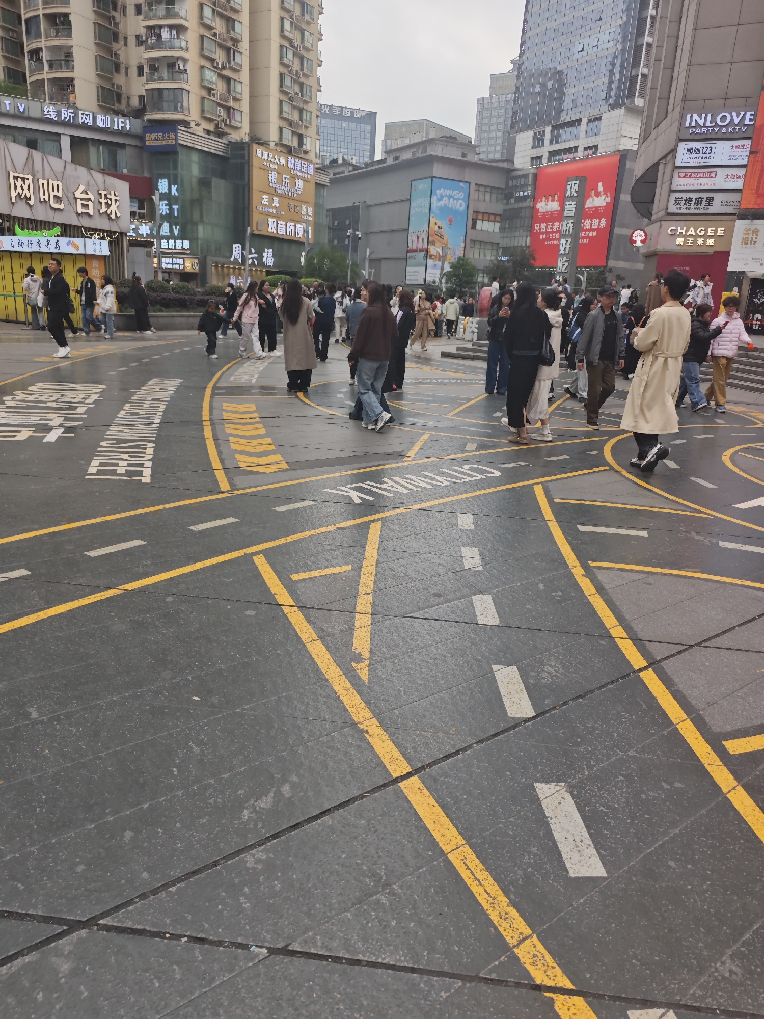 The photograph depicts a bustling pedestrian zone in Liangjiang, China. The ground is paved with grey stone, marked with yellow and white lines resembling roadways. Numerous pedestrians, dressed in diverse clothing, are walking across the marked pavement. Several tall buildings with advertisements and shopfronts form the backdrop of the scene. Signage in Chinese and English, including the names of brands such as "CHAGEE", "INLOVE", and "MINISO", are visible on the buildings. Overall, the image portrays a busy commercial intersection.