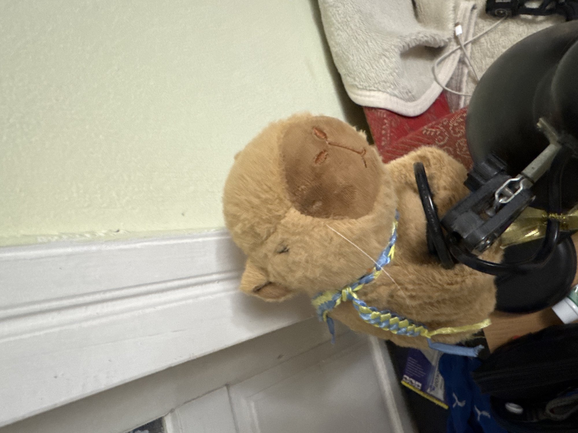 A light brown, plush stuffed capybara toy is positioned centrally in the foreground. The toy is adorned with a braided blue and yellow ribbon around its neck. To the left of the capybara is a white, ornate door frame. Behind the capybara, a red patterned fabric is visible, partially covered by a light gray textured blanket or towel. In the lower foreground, parts of a black object, possibly a piece of machinery or a large accessory, are present. Scattered items, including what appears to be a blue and white packet and a white bottle, are visible on the floor to the left of the capybara. The background wall is painted a light yellow-green color. The lighting in the scene suggests an indoor environment, possibly a room.
