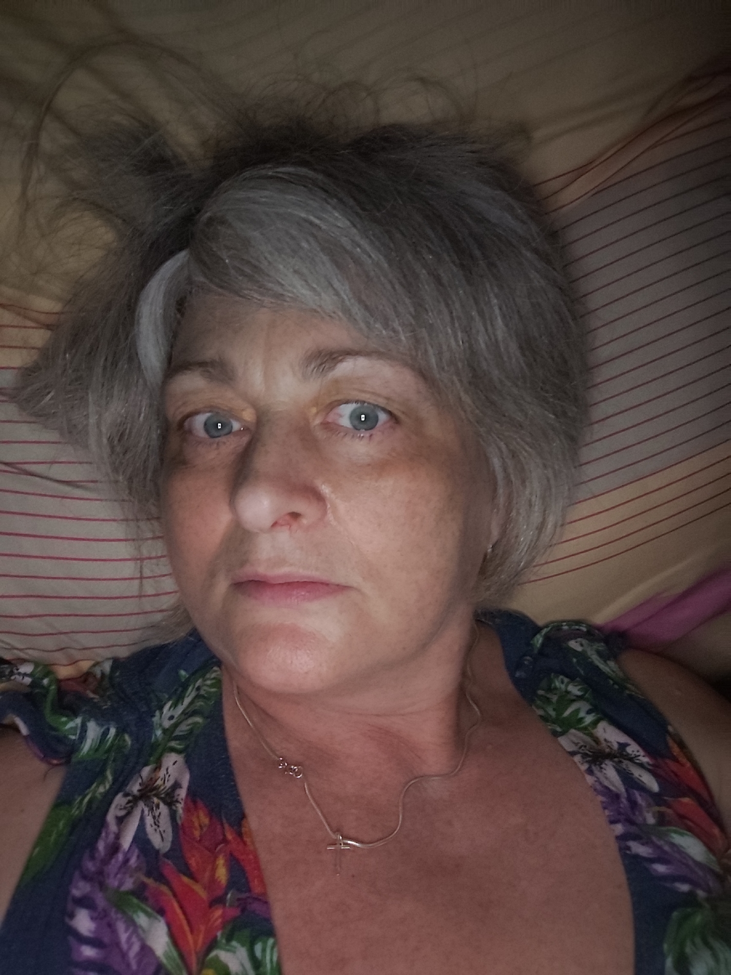 A woman with gray hair and blue eyes is lying down, possibly in bed, as indicated by the striped pillow behind her. She is looking at the camera. She is wearing a floral-print top and a silver necklace with a cross pendant. The lighting suggests the photo was taken indoors, possibly at night, with the camera being held close to her face, indicating a selfie. There is no visible indication of the location in Krasnodar, Russia.