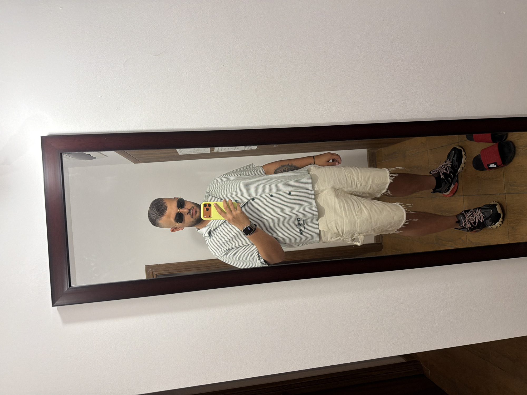 In a hotel room in Nesebar, Bulgaria, a man with a short buzz cut and sunglasses takes a mirror selfie. He is wearing a vertically striped short-sleeved button-down shirt, off-white cargo shorts with frayed hems, black athletic sneakers with pink laces, and a black smartwatch on his left wrist. He is holding a bright yellow smartphone in his right hand, with which he is taking the picture. To the left of the man, a dark-framed full-length mirror reflects his image and a portion of the room. The wall behind the mirror is painted white. The floor is tiled with a wood-like pattern.
