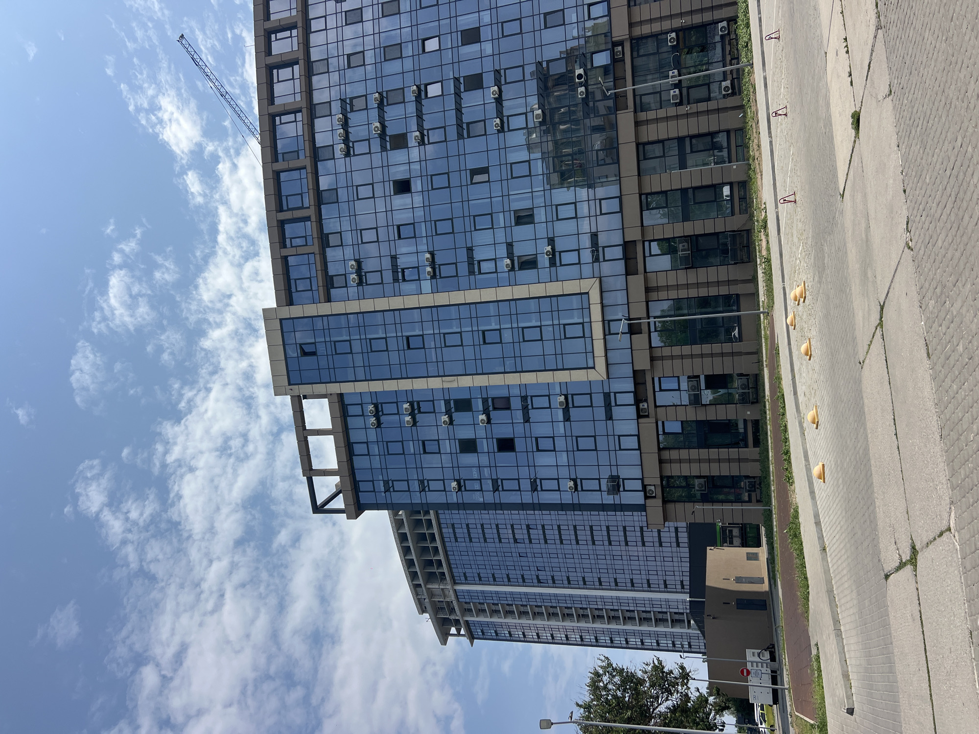 An eye-level, medium-shot, vertical photograph captures a modern, multi-story building with a blue glass facade and brown accents, situated in the city of Fontanka, Ukraine. The building is partially under construction, with the upper floors of an adjacent taller building showing exposed concrete and rebar. Several air conditioning units are visible on the exterior of the glass-fronted building, attached to the facade between windows. A construction crane is visible in the upper right quadrant of the image, extending from behind the building. In the foreground, a gray paved surface with yellow bollards and red parking barriers indicates a parking lot or outdoor area. A small, brown, single-story building with a gray door is located to the left of the main structure, with a traffic sign nearby. Trees and a streetlamp are present to the left of the scene, and the sky above is a bright blue with scattered white clouds. The lighting suggests it is daytime, likely midday or early afternoon. No people are visible in the image.