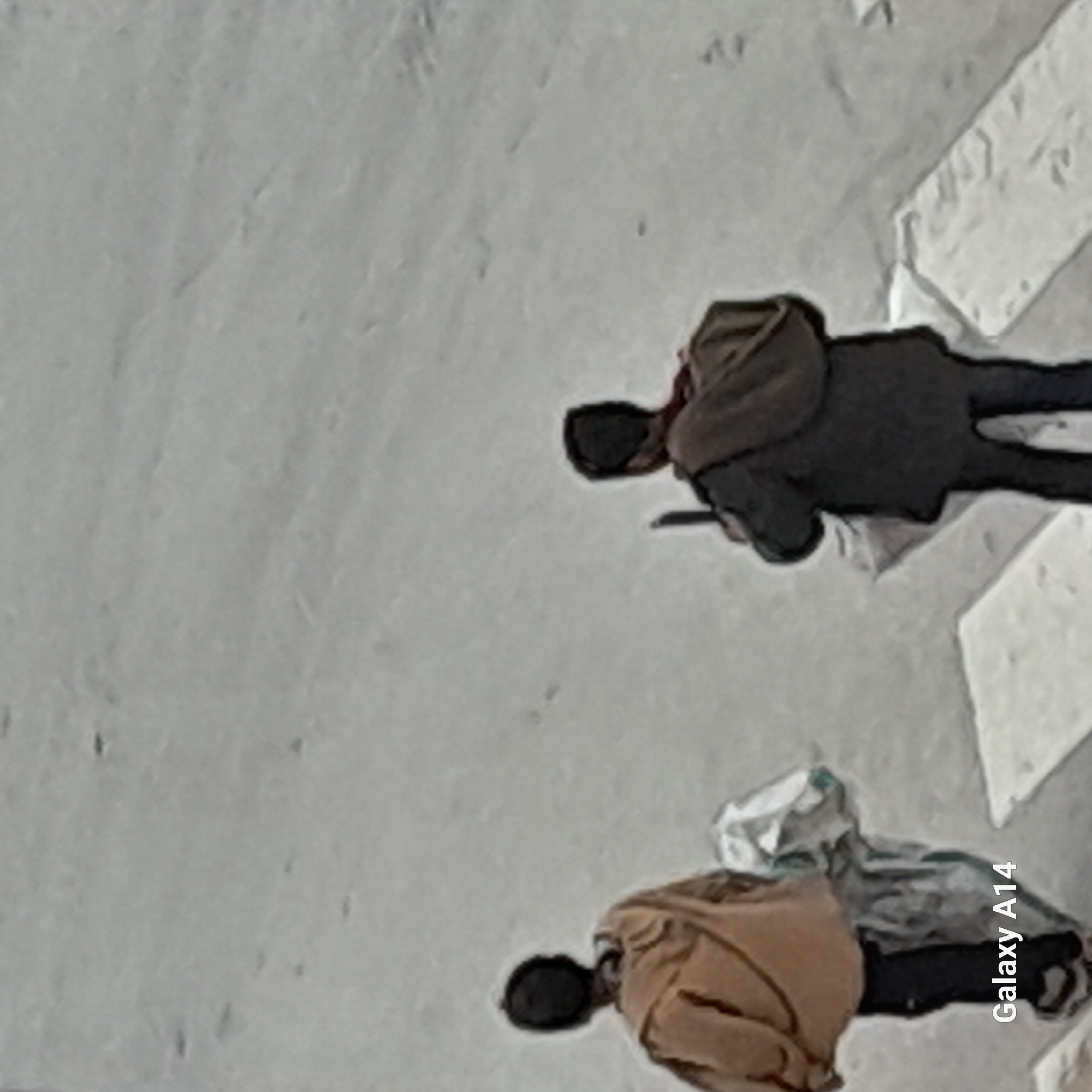 The image shows two people lying on a gray surface in the city of Unknown, Unknown. The person in the lower part of the image is wearing a tan-colored coat and black pants, and has what appears to be a white bag next to them. The person in the upper part of the image is wearing a dark-colored coat and black pants. The background is a gray, slightly textured surface. The text "Galaxy A14" is visible in the lower right corner of the image.