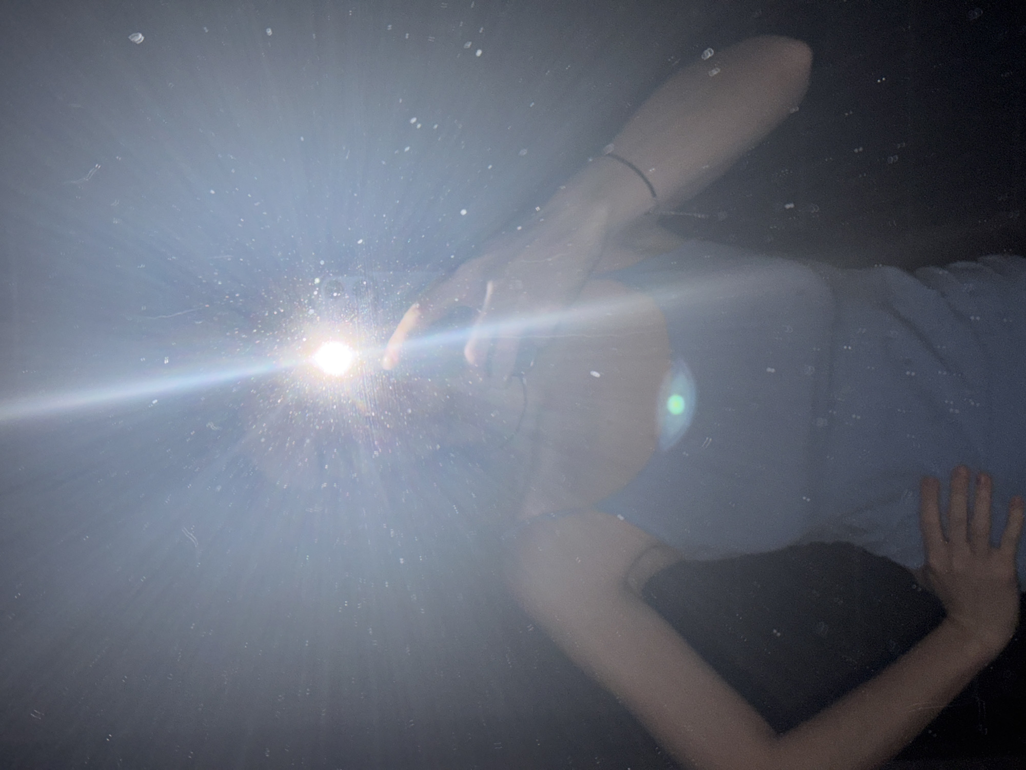 A person is lying down, arm raised to block a bright light. They are wearing a light blue shirt. Their other arm is bent, hand near their torso. The background is dark. The location, Chaikovsky, Russia, cannot be definitively determined from the image.