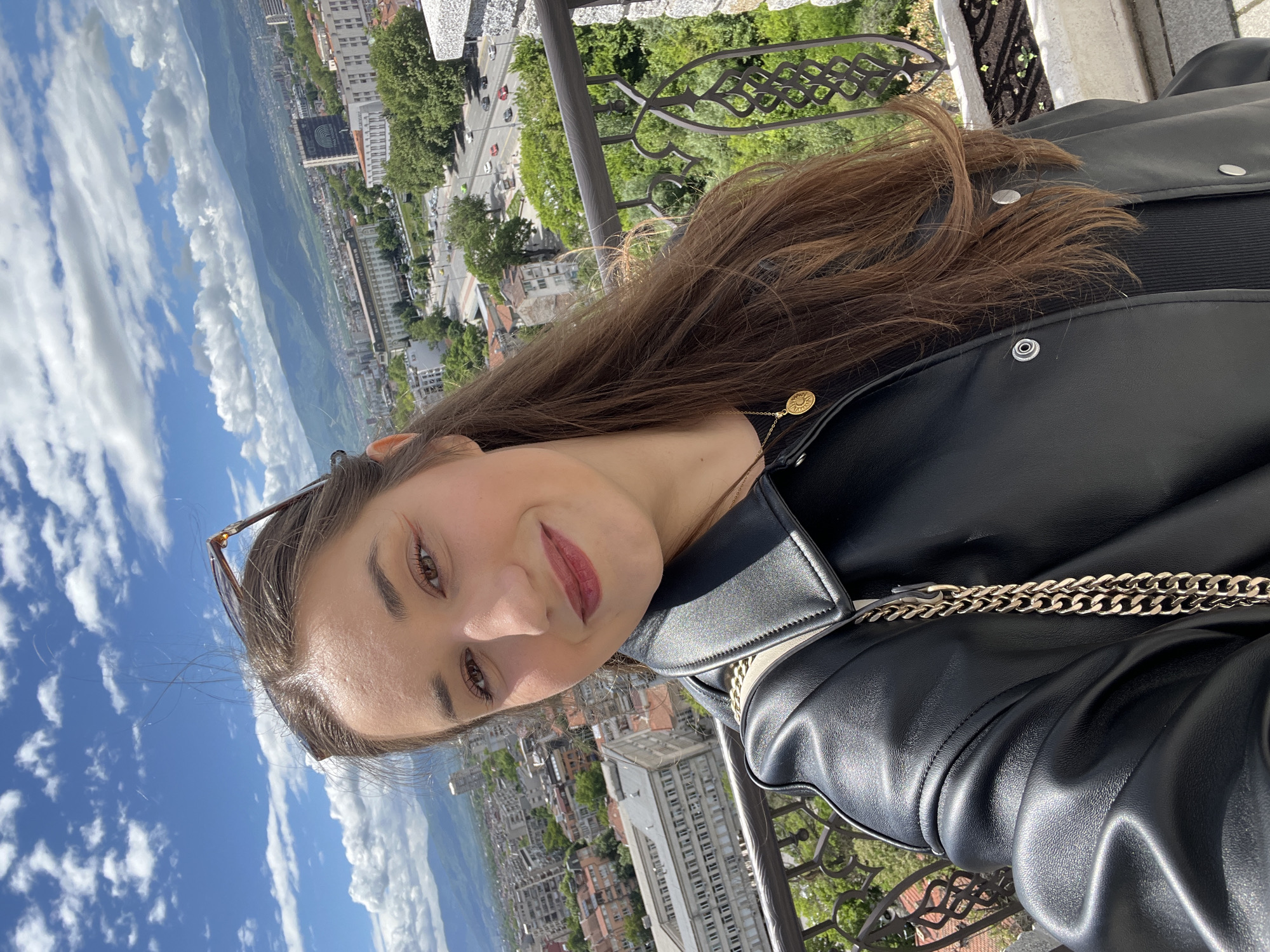 A young woman with brown hair and dark eyes is posing for a selfie. She is wearing a black leather jacket and a black top. She is standing on a balcony with a wrought iron railing overlooking the city of Plovdiv, Bulgaria. In the background, there are buildings, trees, mountains, and a blue sky with white clouds. The city appears to be in a valley between mountain ranges.