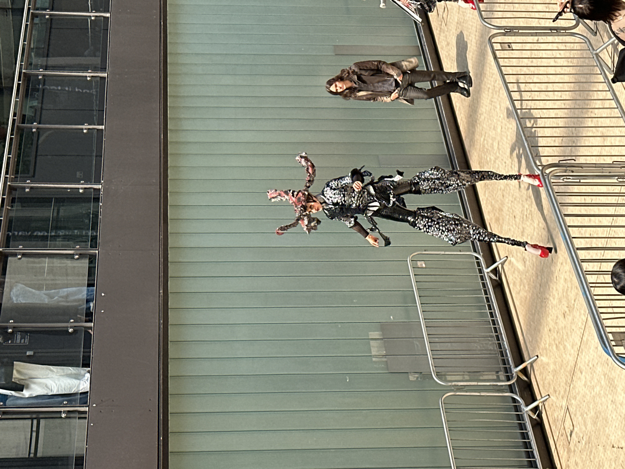 The image depicts a street scene in what appears to be London, United Kingdom, based on architectural styles and street furniture. A person in a black and silver costume with elaborate headwear is posing on the sidewalk behind a barrier. A person in casual clothes (brown jacket and dark trousers) is standing nearby, watching the person in the costume. The background includes a modern building with multiple glass windows and a grey facade. The day is bright and sunny.