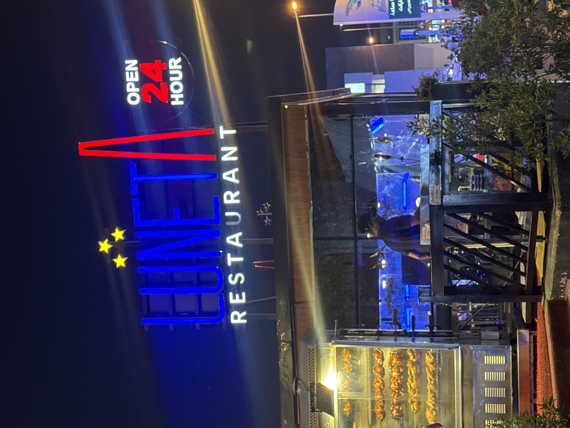 At night, a rotating vertical rotisserie grill holding cooked chickens is displayed in front of UNET Restaurant, which is open 24 hours and located in Dubai, United Arab Emirates. A man in a dark shirt is partially visible through the restaurant's window. The restaurant sign, featuring the name in vertical letters with red and blue neon lights and three yellow stars, is prominently displayed. The scene suggests an urban setting with other buildings visible in the background.