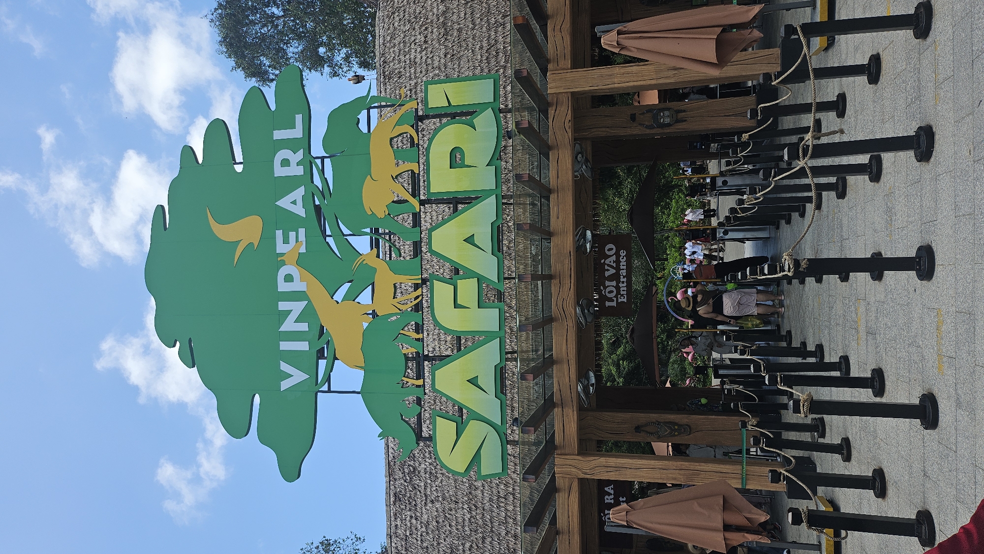 The image shows the entrance to the Vinpearl Safari park in Phú Quốc, Vietnam. A large green sign with "VINPEARL SAFARI" written in bold letters, along with silhouettes of various animals, is prominently displayed above the entrance. The entrance itself is a wooden structure with a thatched roof, and queues of people are visible, suggesting it is a popular attraction. The overall scene is bright and sunny, with a clear blue sky and some clouds.