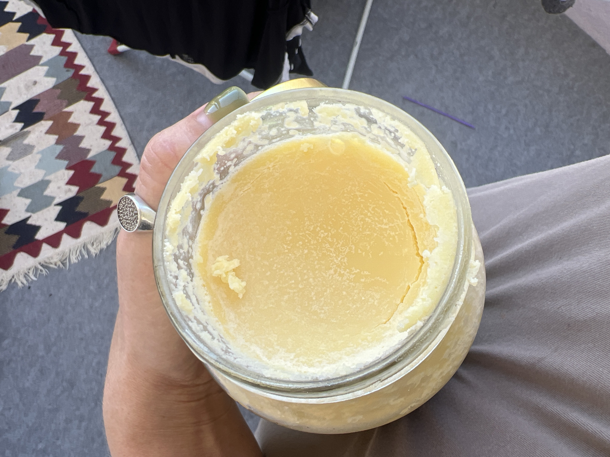 The image shows a close-up, overhead view of a person's hand holding a large glass jar filled with a yellow, semi-solid substance, likely ghee or clarified butter. The jar has a metal lid that is partially visible at the top. The person is wearing a silver ring with small, dark stones on their index finger, and their fingernails appear to be painted with a metallic green polish. The background is out of focus and suggests an indoor setting with a patterned rug and a dark fabric, possibly clothing, to the right. A thin, purple object, possibly a straw or a small stick, is lying on the gray surface in the lower right portion of the image. The overall lighting is natural, suggesting daylight. The image is cropped such that the exact location within Novosibirsk, Russia, cannot be determined.