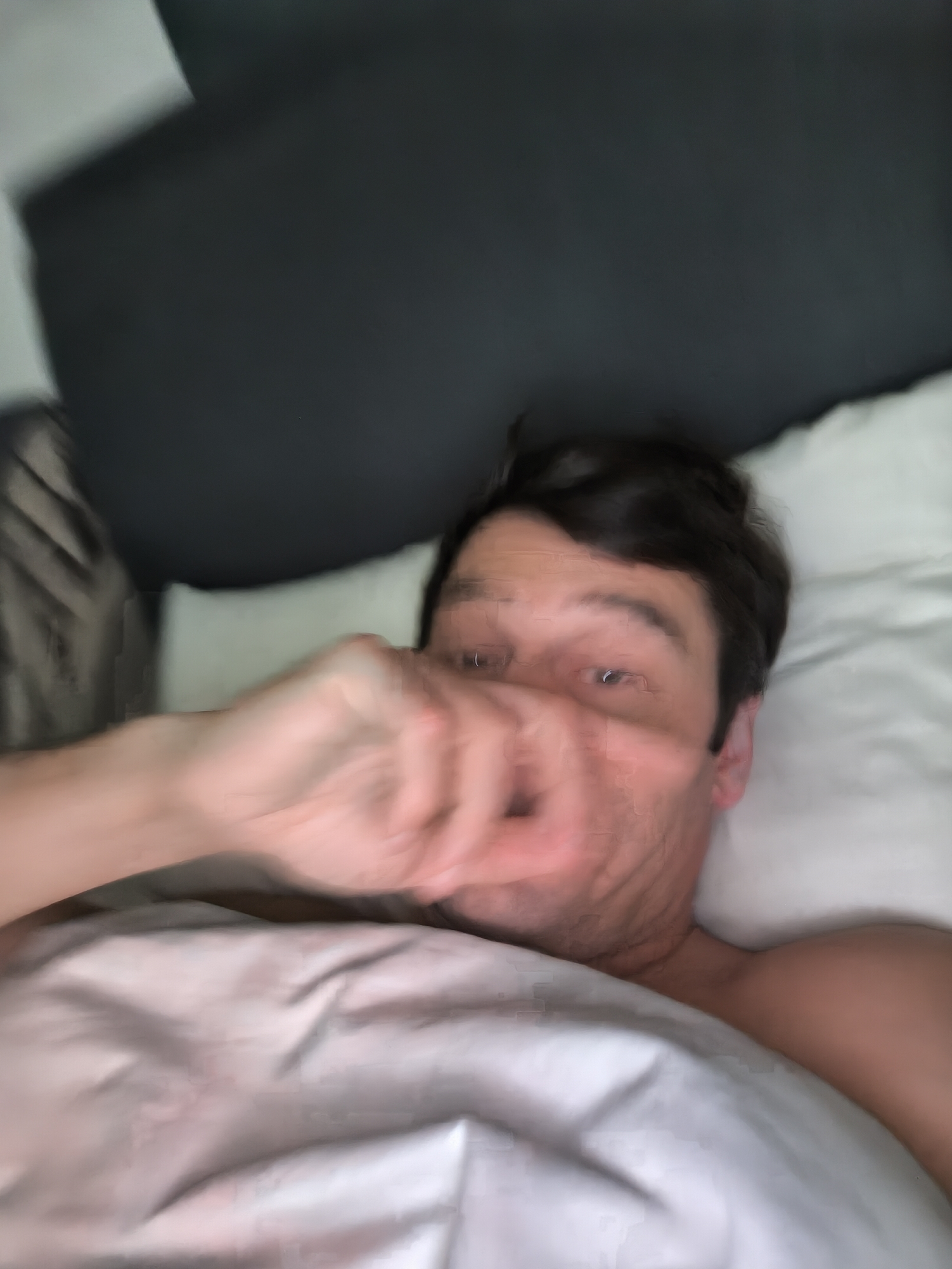 A blurry image shows a man lying in bed. He is partially covered by a white blanket and has a white pillow under his head. He is shirtless and his dark hair is visible. His hand is raised towards his face. A dark headboard is visible behind the pillow. The location is not specified and no information suggests it is Moscow, Russia.