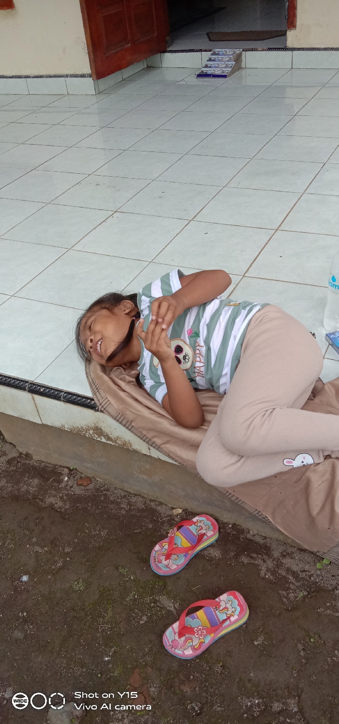 The scene depicts an outdoor tiled patio in Ranugedang, Indonesia, bordering a building and a dirt ground area. A single individual, appearing to be a child, is lying on their left side on a light brown patterned blanket covering the white square tiles. The individual wears a light green and white striped t-shirt with a 'HAPPY' graphic and beige ribbed pants featuring a white bunny emblem on the left leg. Their dark hair is pulled back, and their face is turned upward with an open-mouthed, smiling or laughing expression, eyes open. Their hands are clasped near their chest, seemingly interacting with a small, indistinct object. Adjacent to the individual on the tiled floor, a clear plastic water bottle with a blue label and a small blue rectangular object are visible. The building entrance in the background features a partially open dark reddish-brown wooden double door, a dark doormat inside, and a low, blue and white tiled ramp leading into the doorway. Immediately in front of the tiled patio, a lower strip of dark, moist earth with minimal vegetation contains a pair of children's flip-flops; these are pink/red with colorful stripes, unicorn designs, and red straps with flower accents.