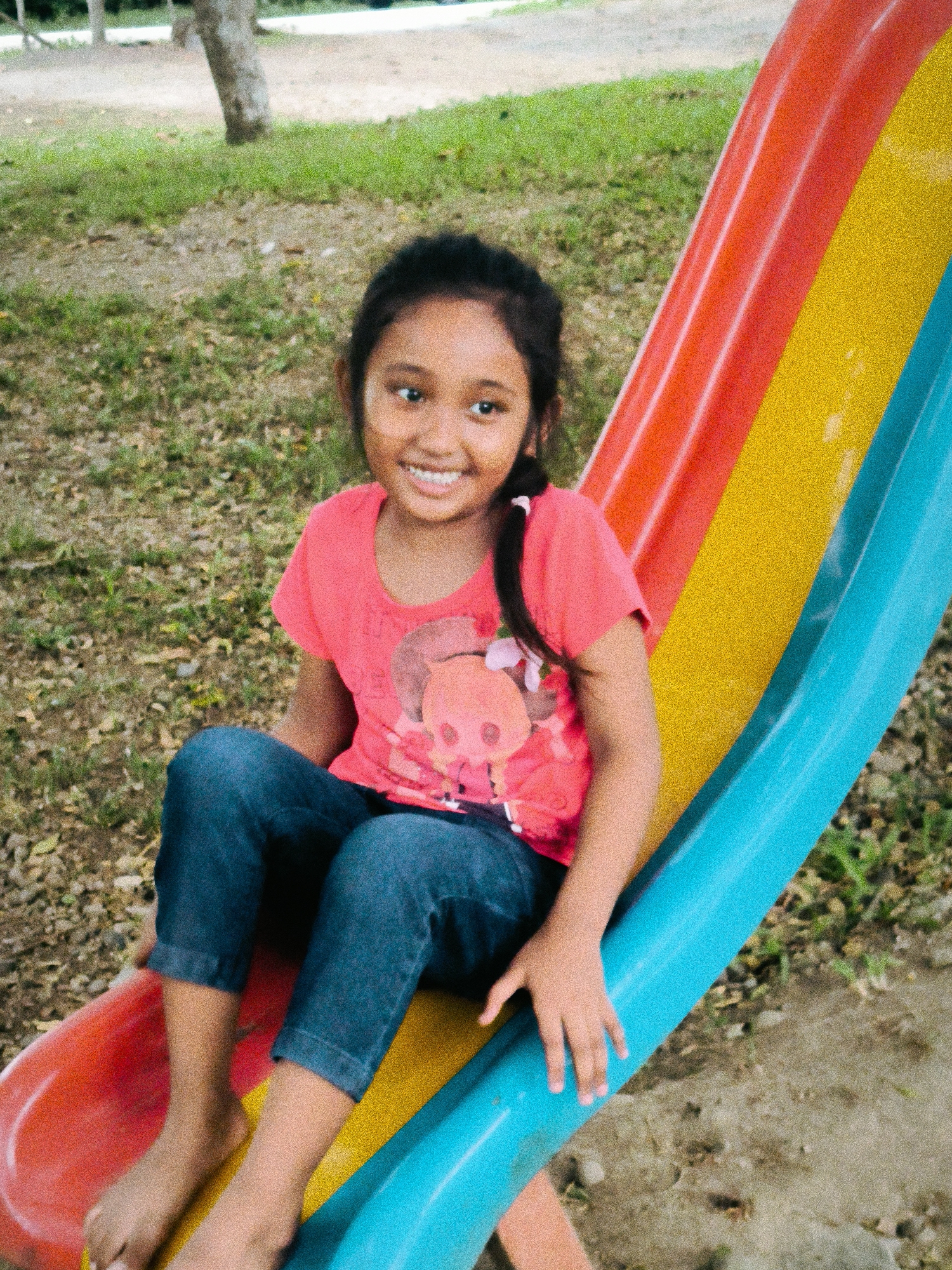 A young girl is sitting on a colorful playground slide outdoors. The slide features distinct red, yellow, and blue sections, with the girl positioned on the red and yellow parts. She is smiling broadly and looking towards the camera, appearing happy and playful.
The girl has dark hair, partially tied back with a pink hair tie, and is wearing a coral or pink t-shirt with a graphic design of what appears to be a cartoon pig or similar animal character with floral elements. She is also wearing blue denim jeans or capris and is barefoot, with her feet resting on the yellow section of the slide. Her left hand is resting on the blue section of the slide.
In the background, there is a grassy area with some sparse vegetation and bare ground, along with the trunk of a tree. The lighting suggests it is daytime, possibly morning or afternoon, with clear weather. The image has a somewhat grainy texture. The scene depicts a child enjoying an outdoor play area in Lamtamot, Indonesia. No visible text is discernible in the image.