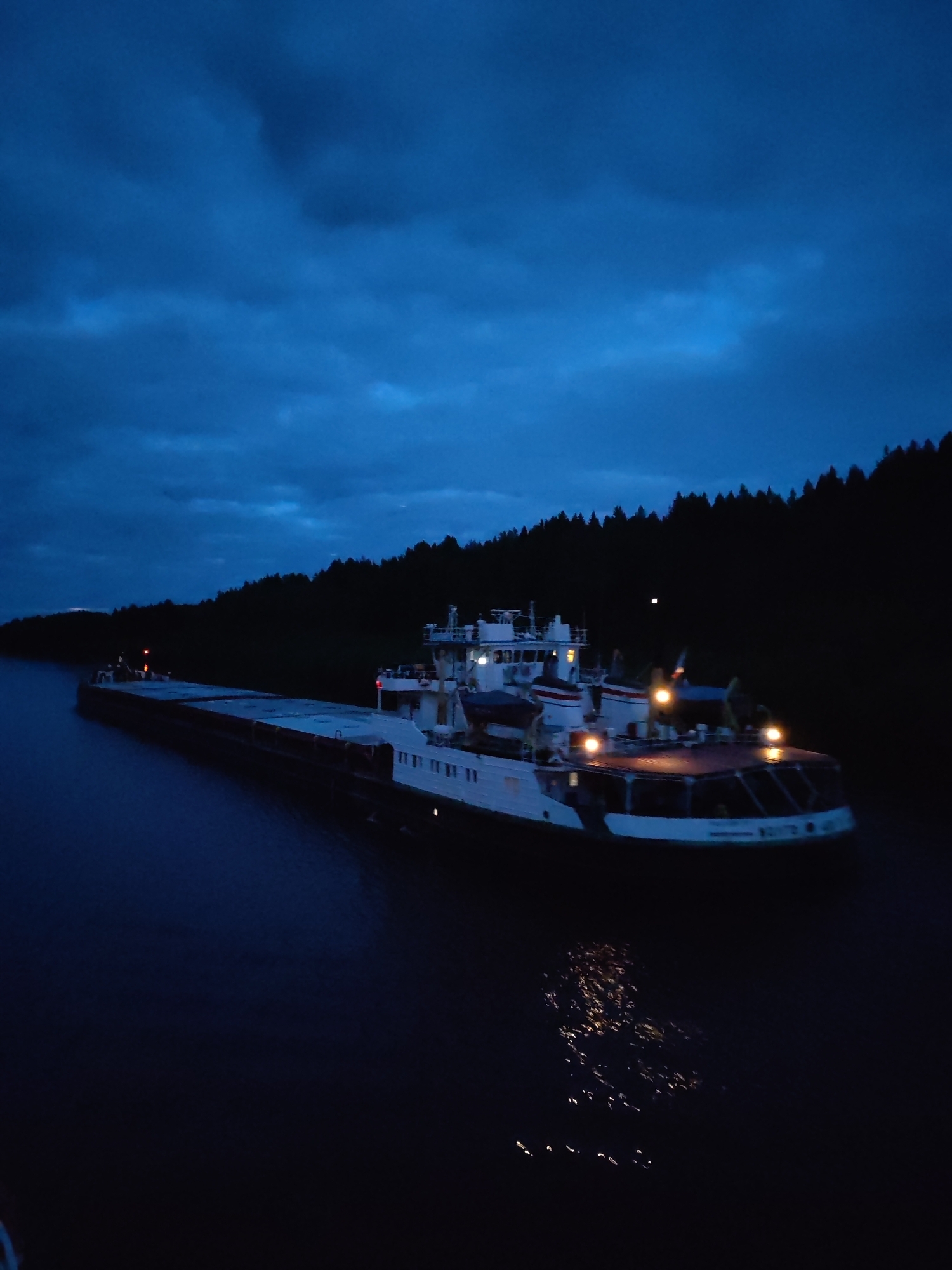 A barge navigates the water near Старое Петровское, Russia, at dusk or night. The sky above is filled with dark, moody clouds. The barge, well-lit, is the main focal point against the dark water. Behind the barge, a tree-covered shoreline is visible.