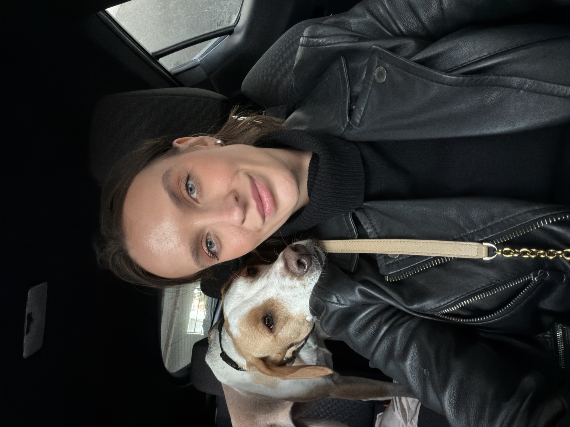 The image depicts an adult female and a dog inside a vehicle. The female, positioned in the foreground, has light skin, blue eyes, and brown hair, and is wearing a black turtleneck under a black leather jacket. A beige bag strap with a gold chain is visible across her torso. She is looking directly at the camera with a slight smile. To her left and partially behind her, a medium-sized dog with white and tan/light brown patches is visible, wearing a black harness. Its head rests on the female's left shoulder, and it is looking towards the camera. Its front paws are on a light-colored fabric or blanket. The vehicle interior features black fabric seats and a dark headliner. A light fixture or sensor is visible on the ceiling. Blurred exterior views are discernible through the rear and side windows. The scene is located in the city of Budva, Montenegro.