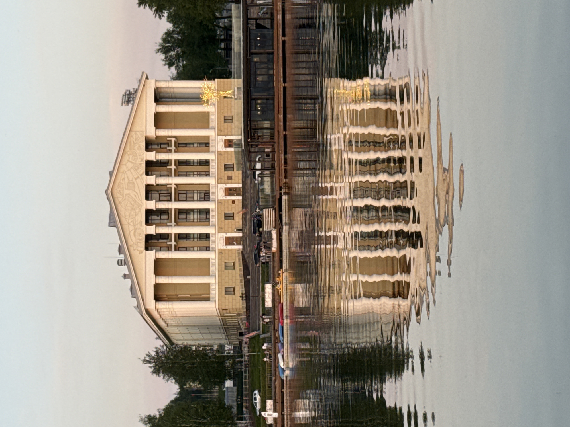 The image shows a building reflected in water. The building has a unique A-frame roof, multiple floors, and a golden star on top. To the right of the building is what appears to be a bridge or walkway. There are trees and greenery on either side of the scene. In the water, the reflection is distorted by ripples. The building is the "Museum of History and Culture of Votkinsk", located in Votkinsk, Russia.
