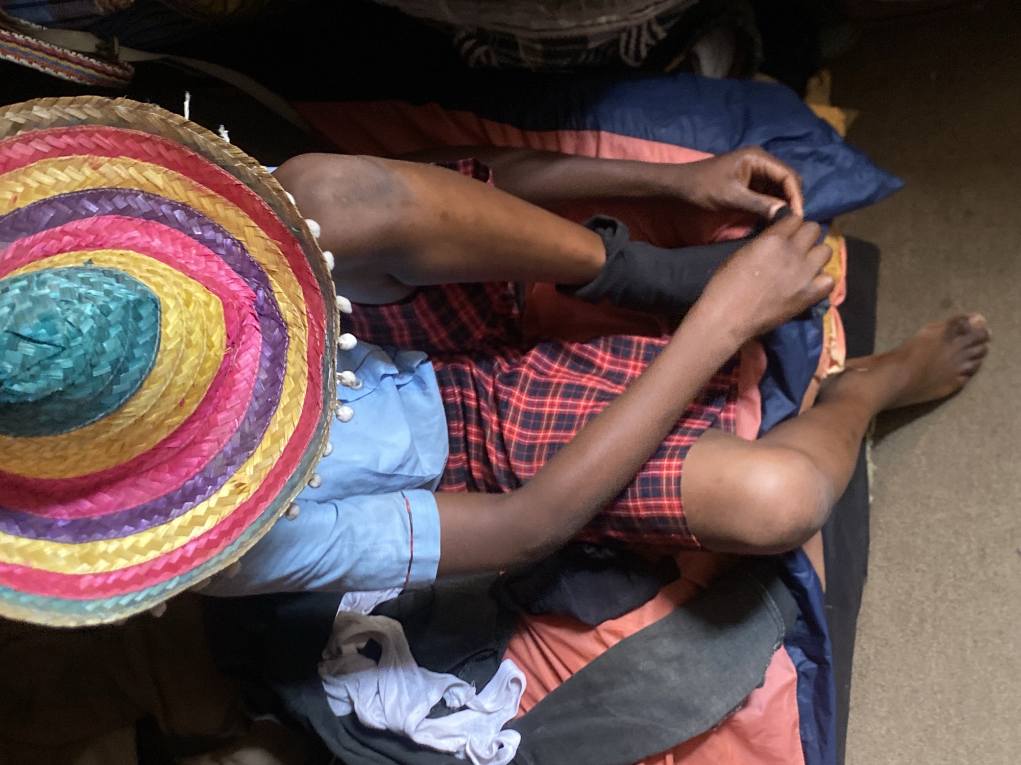 An overhead shot shows a person lying on what appears to be a bed or makeshift sleeping arrangement. The bed consists of blankets and pillows. The person, dressed in a blue shirt and plaid shorts, is putting on a black sock. A sombrero is placed to the side, with its colorful, layered design dominating the left portion of the image. The surrounding environment looks like an indoor space, possibly a room, with personal belongings scattered around. No specific landmarks visible, unable to confirm the location in Bukuru, Nigeria.