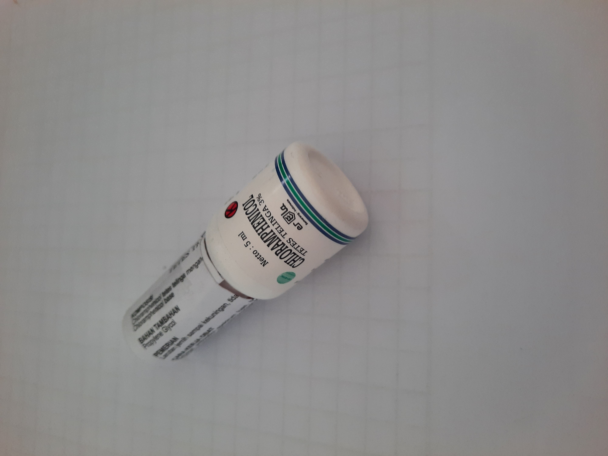 The image shows a small, white bottle of Chloramphenicol ear drops, 3% concentration, with a net volume of 5 ml. The bottle has a label with Indonesian text, including "BAHAN TAMBAHAN" (Additional Ingredients) and "Propylene Glycol". There is also a red circular symbol with a bat silhouette, indicating it is an over-the-counter medication. The bottle is lying on a light-colored, subtly patterned surface that appears to be a table or desk. The lighting is bright, casting a slight shadow from the bottle. The background is out of focus and appears to be a wall or plain surface. There are no people or animals visible in the image. The bottle is the sole subject.
The image does not contain any visual information that would allow for the determination of its location within the city of Lhokseumawe, Indonesia.