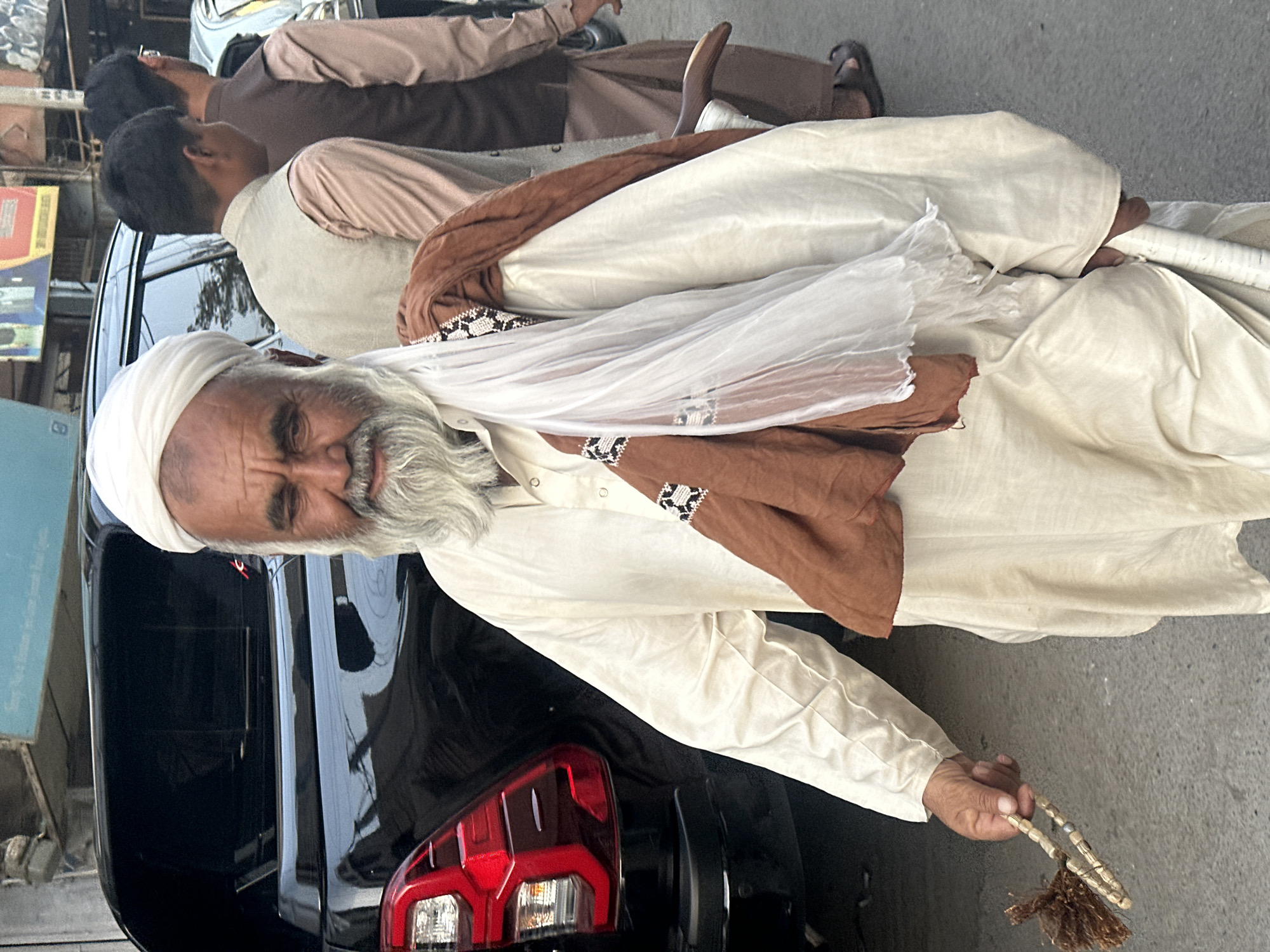 An elderly man with a white turban and a long white beard stands on a paved street in Quetta City Tehsil, Pakistan. He is wearing a loose-fitting cream-colored tunic with embroidered cuffs. A brown cloth is draped over his shoulders, along with a sheer white scarf. In his right hand, he holds a string of beads with a fringed tassel. He is also holding a white cane or stick in his left arm. Behind him, a black SUV is parked, and two other men are visible in the background, one wearing a tan vest over a light pink shirt and the other in a brown vest over a light brown shirt. The environment appears to be an urban setting, possibly a street market or residential area.