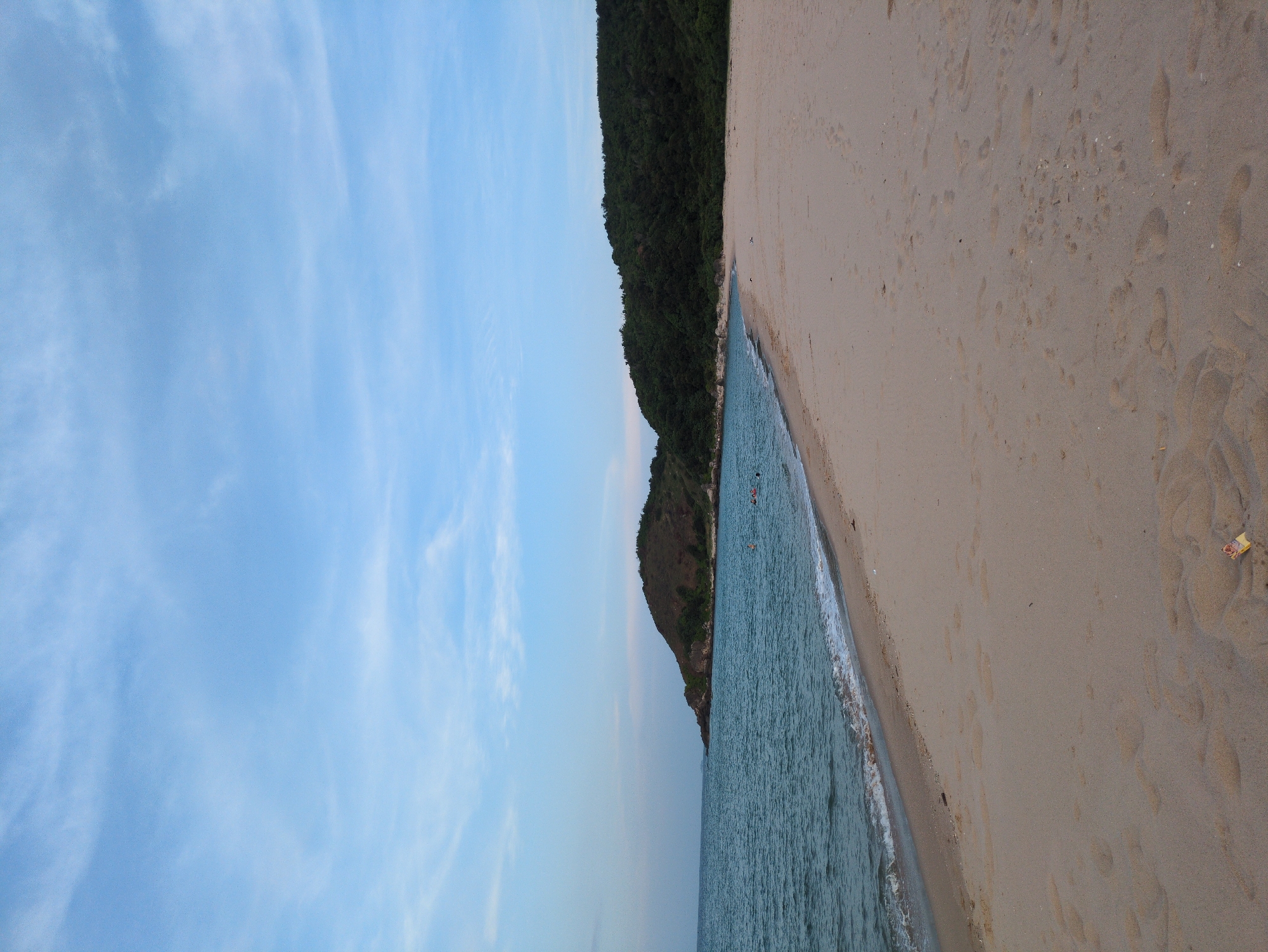 This is a wide-angle, outdoor photograph of a beach in Hoành Sơn Ward, Vietnam, taken from a low perspective. The sky is a pale blue with thin, wispy clouds. The ocean is a muted blue-green, with gentle waves lapping at the shore. The sand on the beach is light brown and has numerous footprints visible, indicating recent activity. In the distance, to the left, a large, tree-covered hill juts out into the sea. To the right, the beach extends further, meeting a dense, green forest that covers another hill. A few people can be seen swimming or wading in the water near the shoreline. There is a small piece of trash, possibly a wrapper, visible on the sand in the foreground. The lighting suggests it is either early morning or late afternoon.