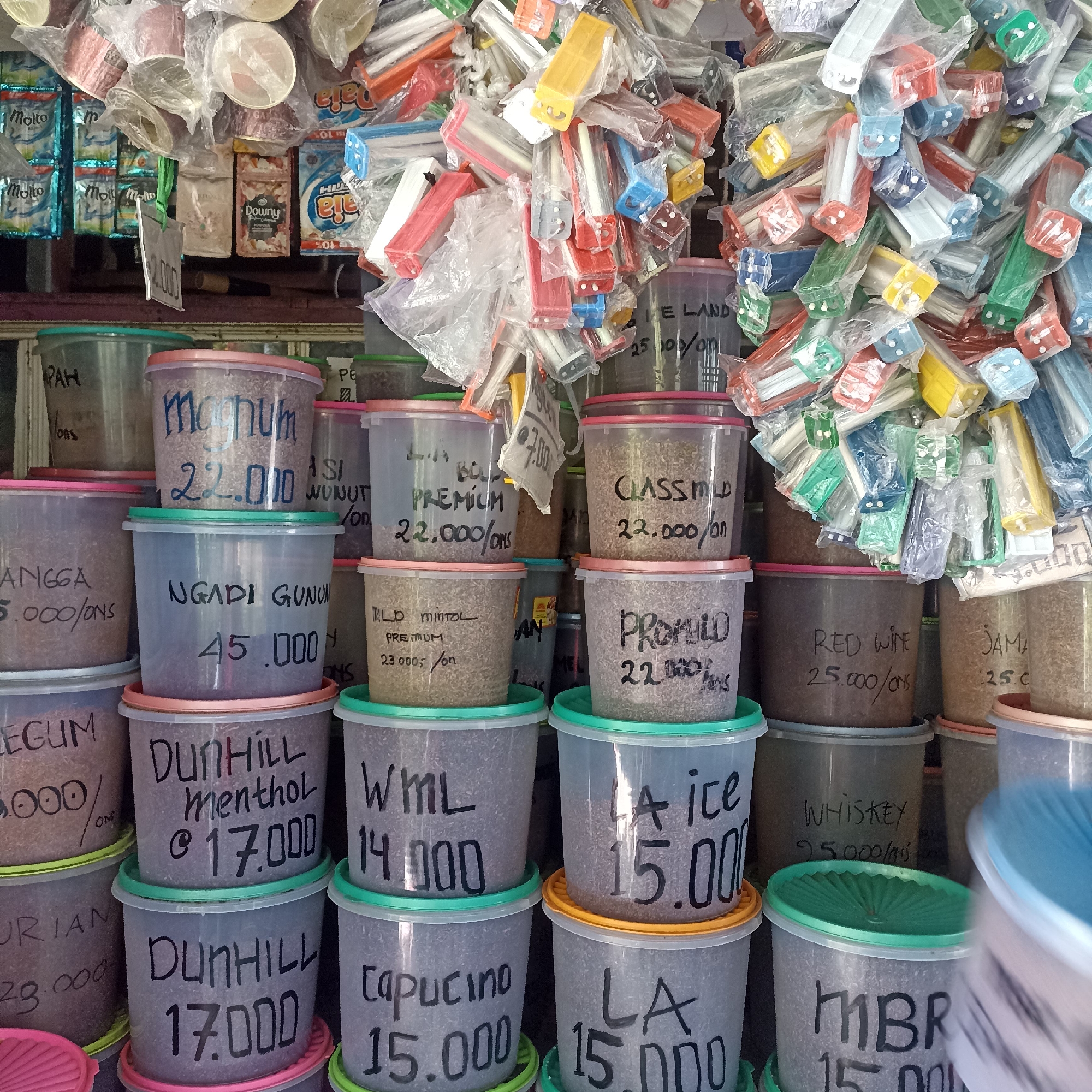 The image shows a close-up, eye-level, outdoor shot of a market stall in Magelang, Indonesia, specializing in loose tobacco. The stall is densely packed with various plastic containers, each filled with a granular substance and labeled with handwritten names of tobacco brands and prices. Brands visible include Magnum, Dunhill, WML, LA Ice, Capuccino, LA, and others. The prices are listed in Indonesian Rupiah, with numbers like "22.000," "45.000," "17.000," "14.000," "15.000," and "25.000" prominently displayed. The containers are stacked and arranged in layers, creating a somewhat chaotic but organized display.
Above the tobacco containers, there are shelves or hanging displays holding various packaged goods, including packets of detergent (e.g., "Molto") and what appear to be cigarette packs. There are also many plastic-wrapped items, some of which are colorful and appear to be toys or accessories, such as brightly colored plastic dispensers that are commonly used for candy or small items.
The lighting suggests it is daytime, with natural light illuminating the scene. The background is slightly blurred, indicating a shallow depth of field. No people are clearly visible in the frame, suggesting the photo was taken from a distance or when the stall was unstaffed. The overall impression is of a busy, local market where individual consumers can purchase tobacco by weight. The specific location in Magelang is not identified visually, but the presence of local products and prices suggests an Indonesian context.