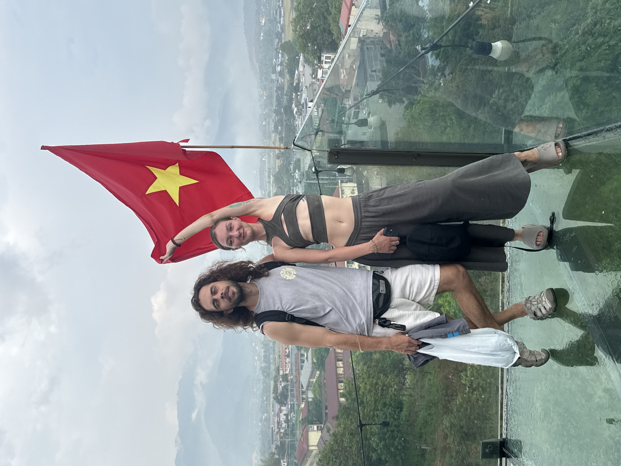 In Nghia Lo Ward, Vietnam, a man and a woman stand on a glass-floored platform with a distant cityscape and mountains visible below. The woman, positioned behind the man, holds a Vietnamese flag to her left. She wears a gray cropped top with cutouts and matching pants, and sandals. The man wears a gray t-shirt, white shorts, and sandals; he holds a white plastic bag. They are both smiling.
