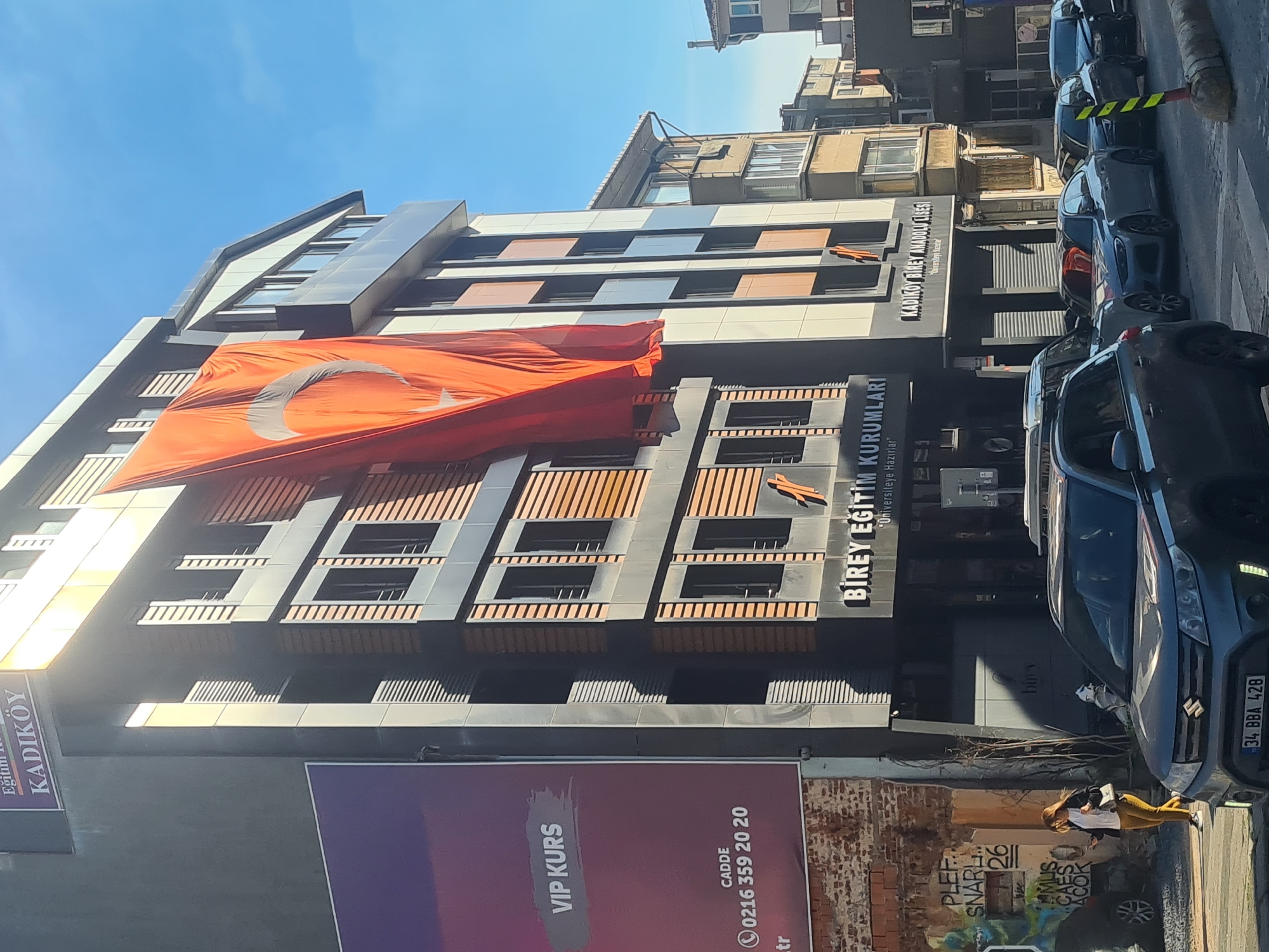 The image depicts a street scene in Kadıköy, Turkey. The focus is on a modern building adorned with a large Turkish flag. The building houses two educational institutions: Birey Eğitim Kurumları (which prepares students for university) and Kadıköy Birey Anadolu Lisesi (an Anatolian high school). A purple billboard advertising "VIP KURS" with the phone number 0216 359 20 20 stands in the foreground. Several cars are parked along the street. A woman wearing yellow pants is walking on the sidewalk in front of a brick wall with graffiti. The sky is clear and blue.
