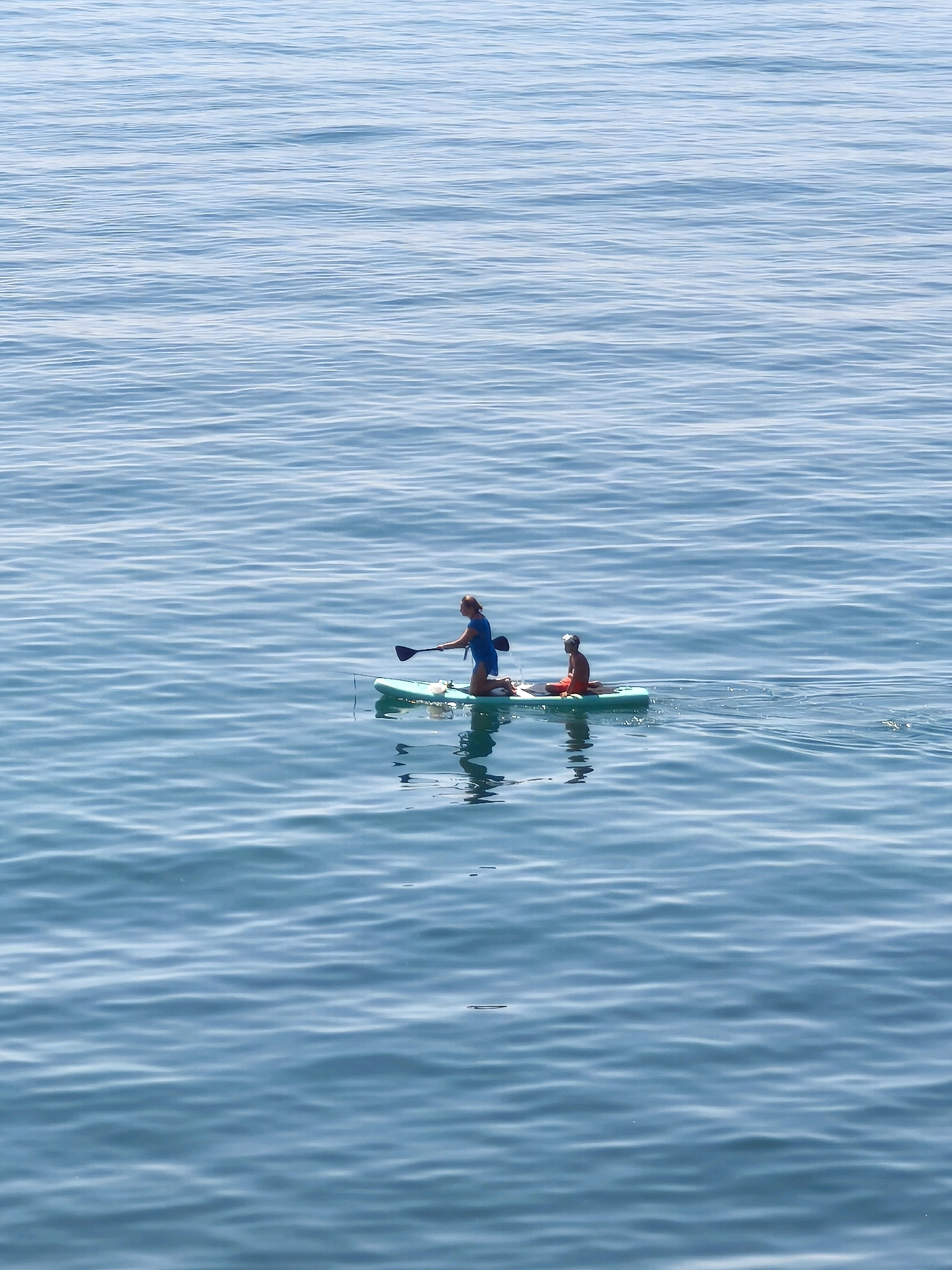 Two people are paddleboarding on a calm body of water. The person in the front is kneeling and paddling with a black paddle. They are wearing a blue top. The person behind them is sitting on the paddleboard and wearing an orange top. The paddleboard is light green and has two people on it. The water is blue and reflects the sky. The scene appears to be outdoors, likely a large lake or the sea, in a location that could be part of Sochi, Russia.