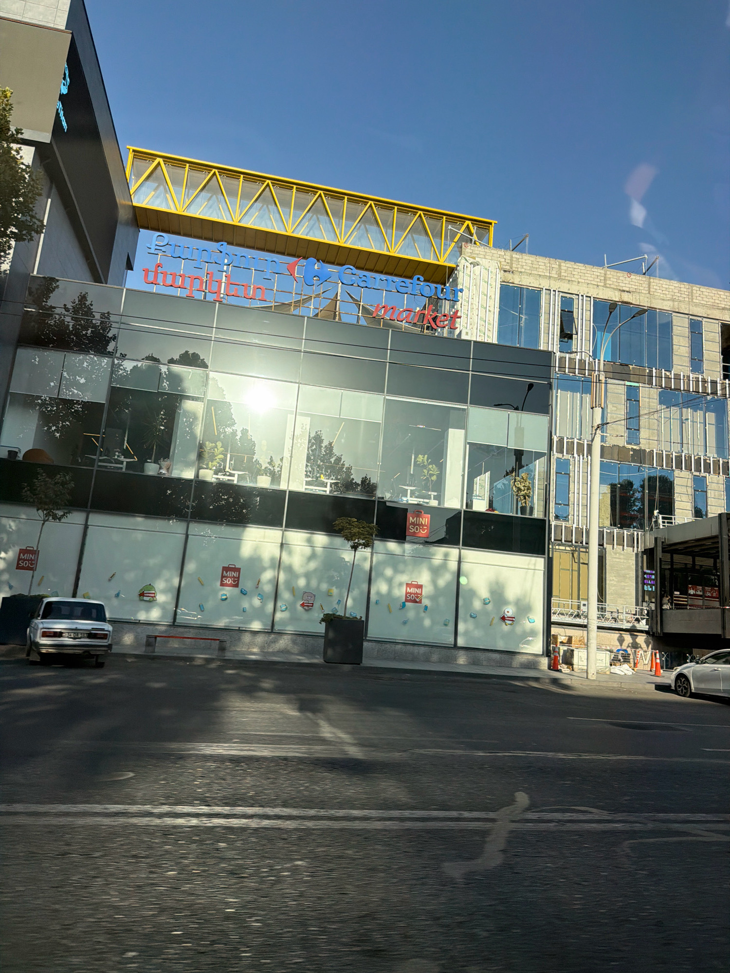 The image shows a sunlit, modern building with a glass facade in Yerevan, Armenia. The building appears to be a commercial establishment, possibly a shopping center or market, as indicated by the prominent signage that reads "Carrefour market" in blue and red lettering. A yellow pedestrian bridge spans across the top of the building, connecting it to another structure. Several cars are visible parked in front of the building, including a white sedan and an older model silver car. The ground in front of the building is paved, and there are trees and planters, suggesting an urban environment. The sky is clear and blue.
