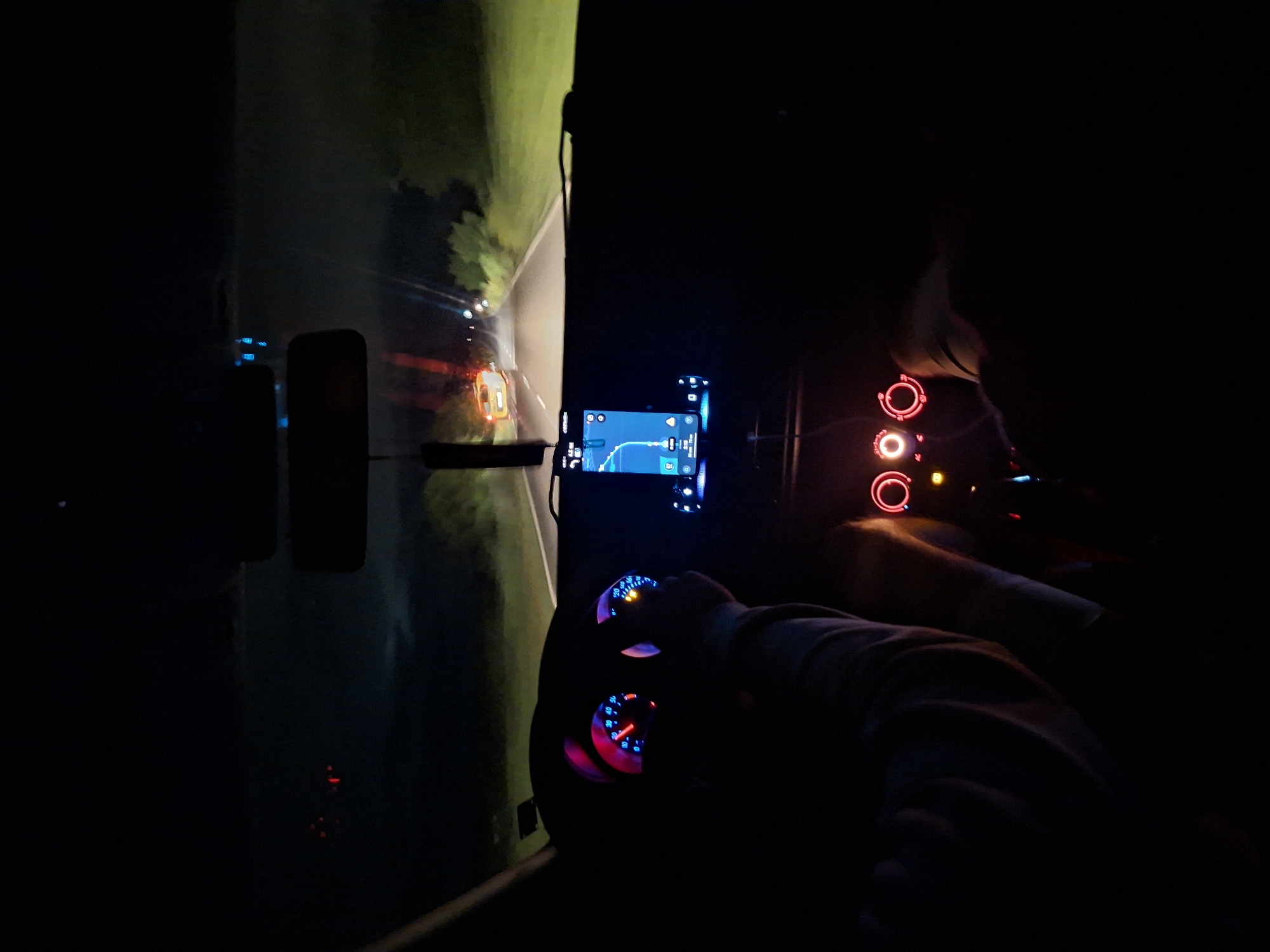 The image is taken from inside a car at night, likely in Kamenar, Bulgaria. The driver's hands are on the steering wheel, which is illuminated by the dashboard lights. A GPS navigation system is displayed on the car's dashboard, showing a route. The road ahead is visible, with the headlights of other cars and streetlights illuminating the dark environment. The scenery outside appears to be rural, with trees lining the road.
