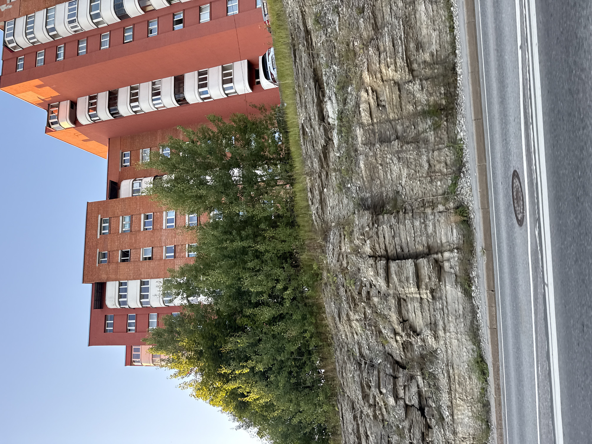 The image shows a multi-story residential building in Tallinn, Estonia, characterized by its prominent orange-red facade and white balcony enclosures. The building appears to be situated on a hillside, with a rocky outcrop and vegetation in the foreground, and a road with lane markings in the lower portion of the frame. A white car is visible parked near the base of the building. The sky is clear and blue, suggesting fair weather. The architecture of the building, with its repeated modular balcony units, is typical of Soviet-era mass housing projects found throughout Eastern Europe.