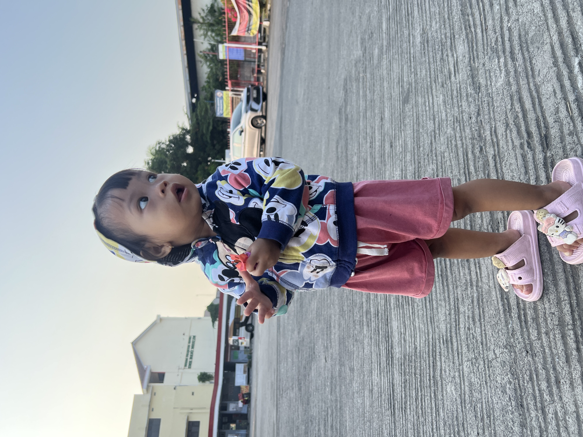 A young child stands on a grey concrete surface, possibly a parking lot or paved area, outdoors. The child is wearing a hooded jacket with a pattern of cartoon characters, pink shorts, and pink sandals adorned with animal charms. The child is holding a small, red object in their right hand. In the background, there are buildings, trees, a car, and a fence. The sky is a pale blue, indicating it might be daytime. The setting appears to be in a developed area, consistent with an urban or suburban location in Kecamatan Pati, Indonesia.