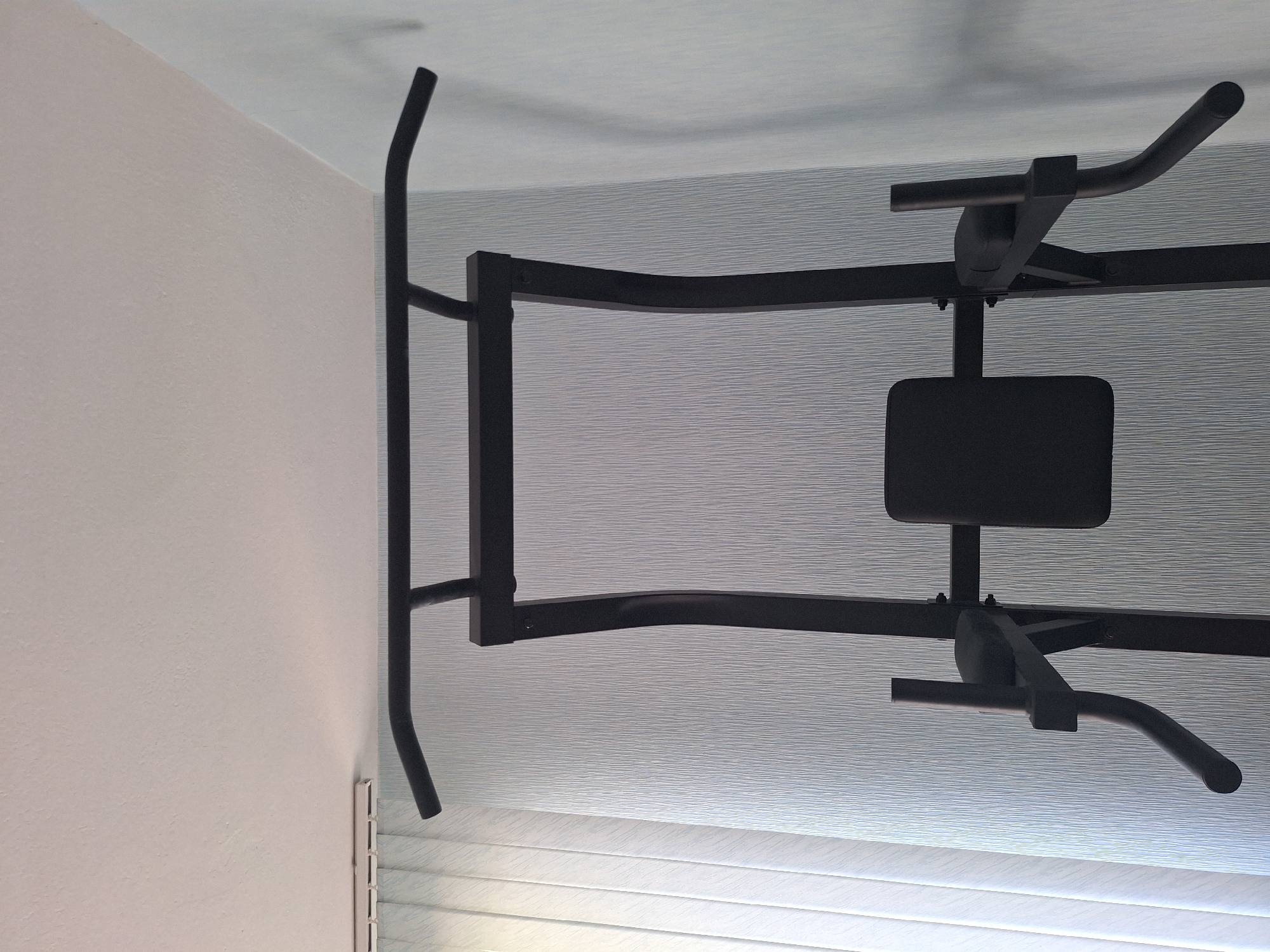 The image shows a black pull-up bar attached to a wall. The pull-up bar is a piece of exercise equipment, and it appears to be a doorway pull-up bar, designed to be mounted in a doorframe. The bar is made of metal and has padded grips for comfort. It is positioned horizontally across the frame of the doorway.
The scene is indoors, likely in a home, given the presence of a wall and what appears to be a doorframe. The wall to the left is painted a light color, possibly white or a very light beige, with a textured finish. The background features a textured surface, possibly wallpaper or the inside of the doorframe, with a horizontally striped pattern. The lighting suggests it is daytime, with the light source coming from the top of the image. There are no people visible in the image. The overall impression is of a functional exercise space within a residential setting in Varna, Bulgaria. There is no visible text in the image.