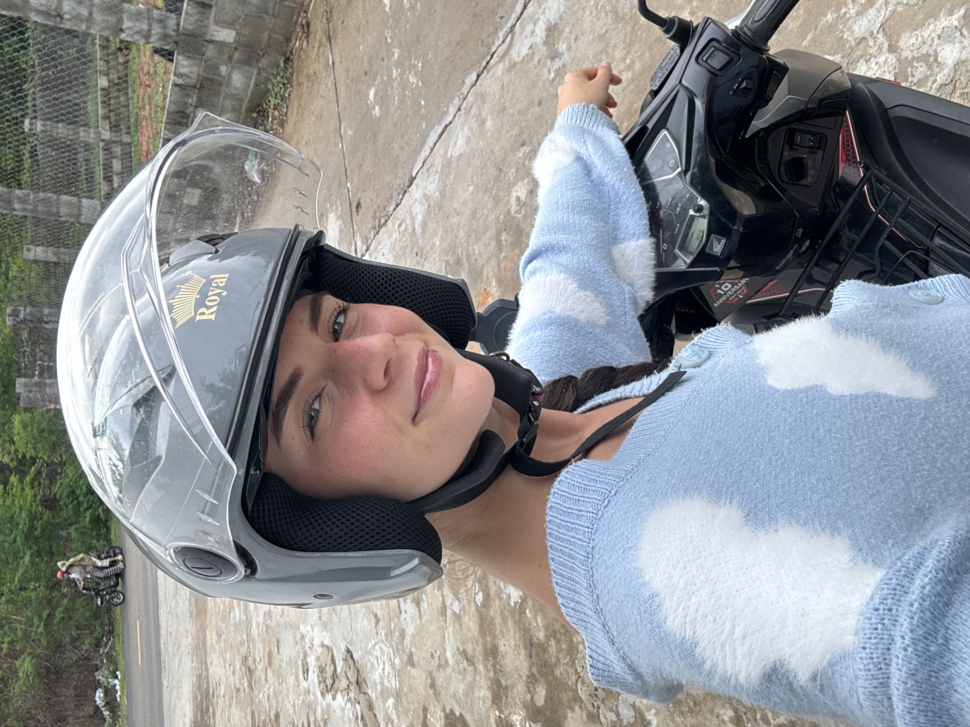 A young woman wearing a gray "Royal" brand motorcycle helmet and a light blue sweater with white cloud patterns is on a motor scooter in Mũi Né, Vietnam. She is positioned on the right side of the frame, her face angled toward the camera. Part of the scooter's black dashboard is visible in the upper right of the image. In the background to the left, there is a glimpse of the road and a distant motorcycle, and a concrete structure sits in the upper left. The ground appears to be a textured, concrete-like surface.