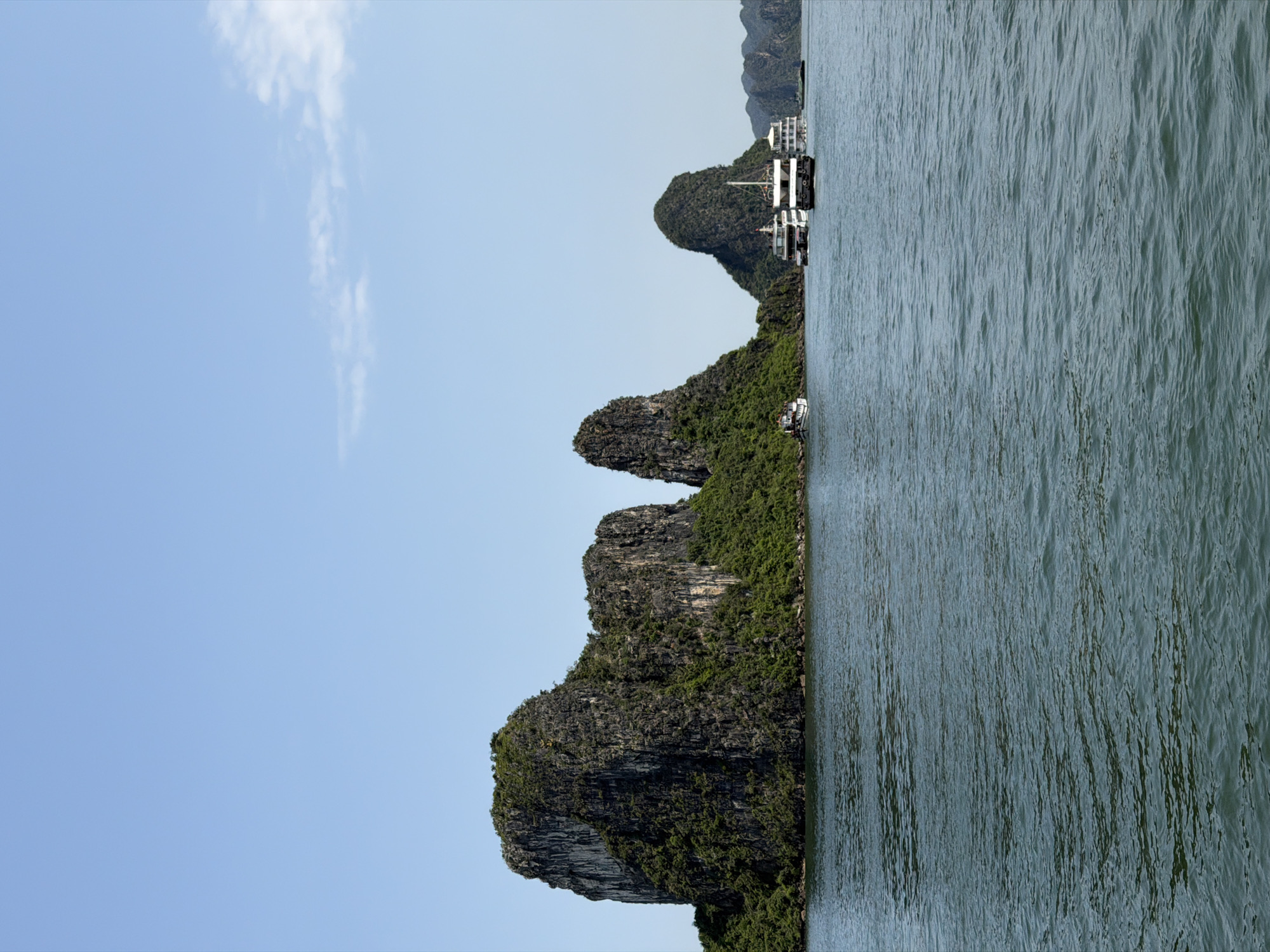 This landscape photograph, presented horizontally, captures a serene daytime view of a bay featuring distinctive limestone karst formations. Several islands, heavily covered in lush green vegetation, rise dramatically from the calm, blue-green water. The sky above is a clear, bright blue with a few wispy white clouds visible towards the left side of the frame.
In the middle distance, nestled between two of the larger islands, are two multi-deck boats, likely cruise ships or tour vessels, appearing to be anchored or docked close to the shore. One boat is prominently featured, showcasing its multi-level white structure. A smaller, single-deck boat is also visible closer to the shoreline of the island in the foreground. The water exhibits gentle ripples, reflecting the clear sky and surrounding scenery.
The scene suggests a tranquil moment in a picturesque natural setting. The bright, even lighting indicates it is likely morning or early afternoon, under fair weather conditions. The unique geological features strongly identify the location as Ha Long Bay, specifically consistent with the area around Tuan Chau Ward, Vietnam. No discernible text or individuals are visible in the image.