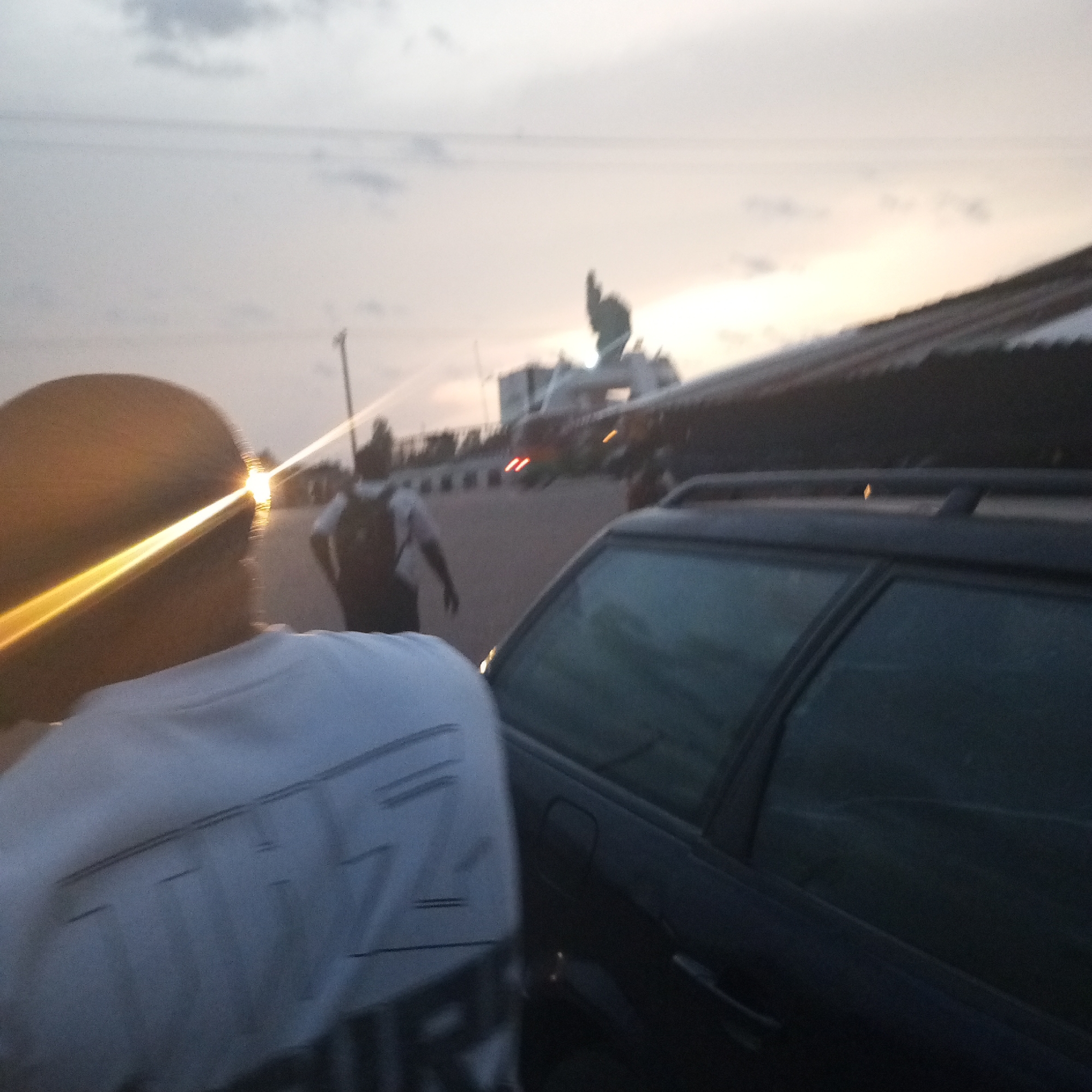 The image depicts an outdoor scene with several subjects. In the foreground, the back of a person wearing a white t-shirt is visible, with a bright reflection shining off the top of their head. To the right, a dark-colored station wagon is partially visible. In the middle ground, another person wearing a white shirt and carrying a backpack is walking away from the camera. In the background, there is a building or structure, possibly a gate, with some details obscured by the bright light. The sky is overcast. The location is in the city of Du, Nigeria.