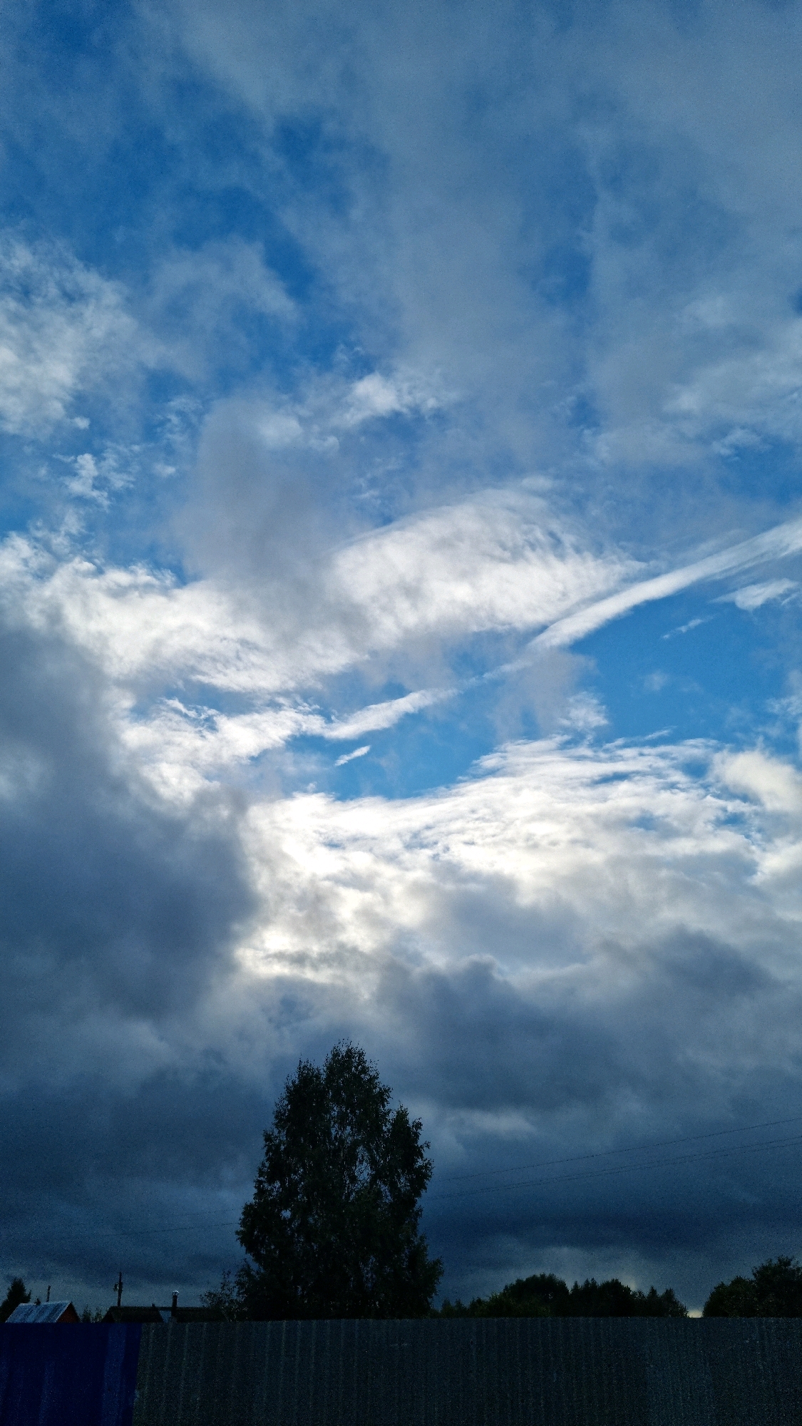 The image shows a cloudy sky over a rural landscape in Vysokoe, Russia. The sky is filled with a mix of dark storm clouds and lighter, wispy clouds, with patches of blue sky visible. Sunlight breaks through in some areas, illuminating the clouds. In the foreground, a tall, leafy tree stands silhouetted against the sky. Below the tree, a dark, corrugated metal fence runs horizontally across the bottom of the frame. Behind the fence and the tree, a line of dark trees forms the horizon, with the roofs of a few small buildings visible amongst them. Power lines are also visible stretching across the middle ground. The overall impression is of an overcast day with potential for rain.