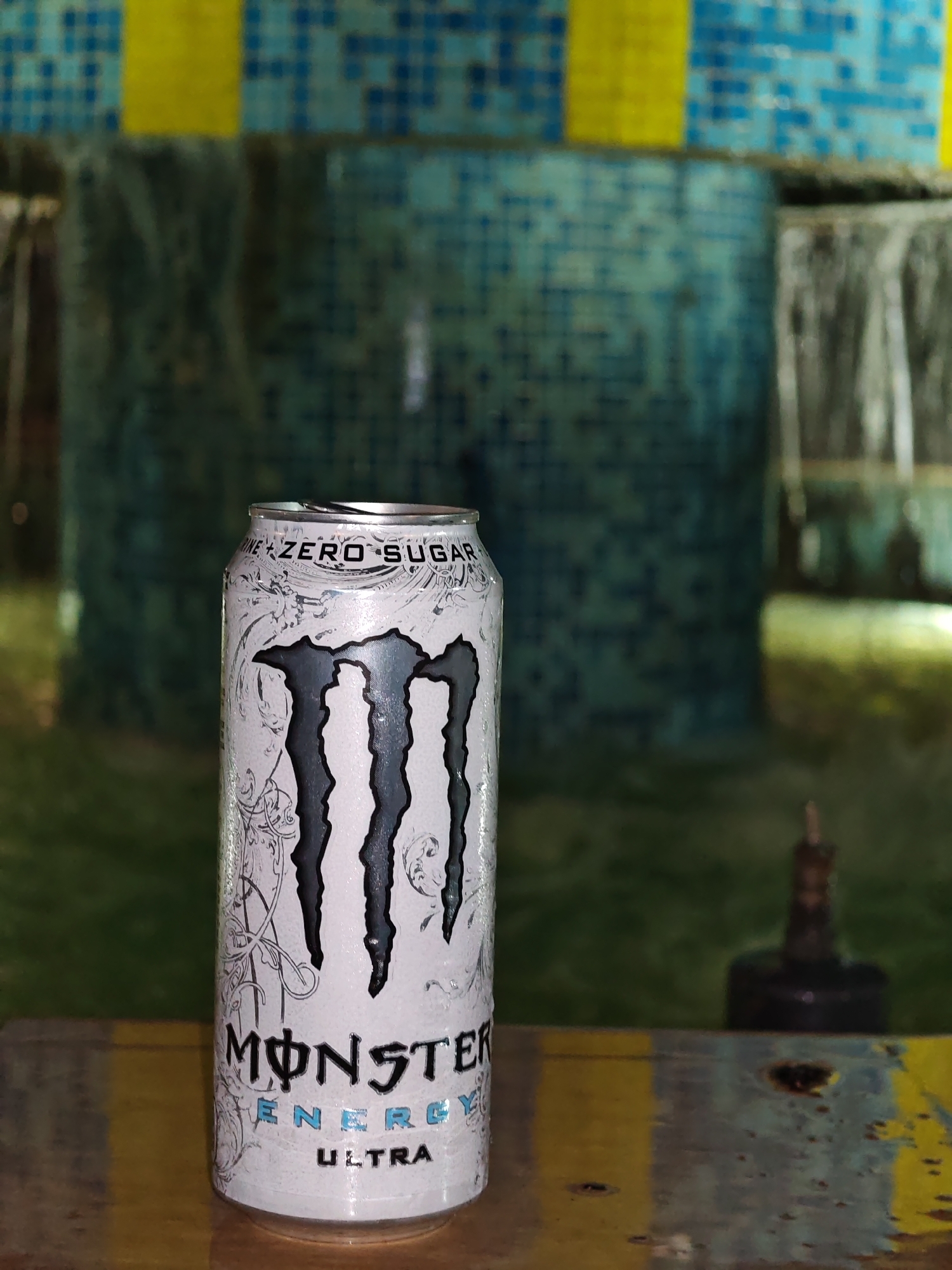 A condensation-covered white Monster Energy Ultra can, labeled "ZERO SUGAR" and "MONSTER ENERGY ULTRA," stands slightly left of center on a worn, multi-colored wooden or metallic surface with yellow and dark blue-green sections. The can's tab is partially opened. The background is significantly blurred, depicting a tiled water feature or fountain. The upper background features blue mosaic tiles interspersed with yellow blocks. A continuous sheet of water forms a waterfall on the right side of the background. A dark, indistinct object with a thin vertical protrusion is visible in the bottom-right background. No individuals are present. The scene is situated in Veliki Preslav, Bulgaria.