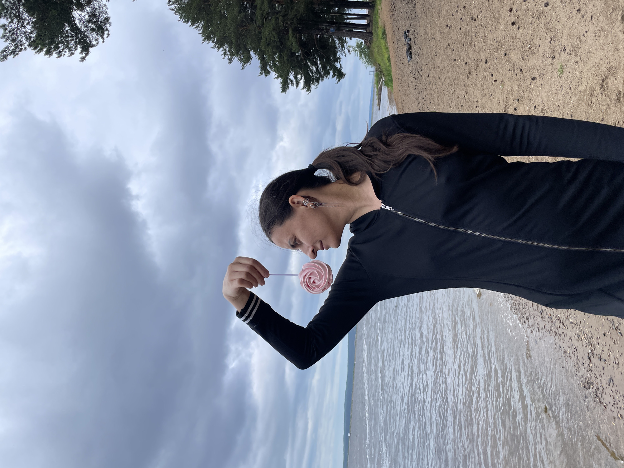 A woman stands on a sandy beach next to a body of water, holding a pink, swirled lollipop above her head. She is wearing a black zippered jacket with white stripes on the cuffs. The sky is overcast with thick clouds. Pine trees are visible in the background to the right, and a distant shoreline can be seen across the water. The scene is located on the bank of the Kama River in the city of Chaikovsky, Russia.