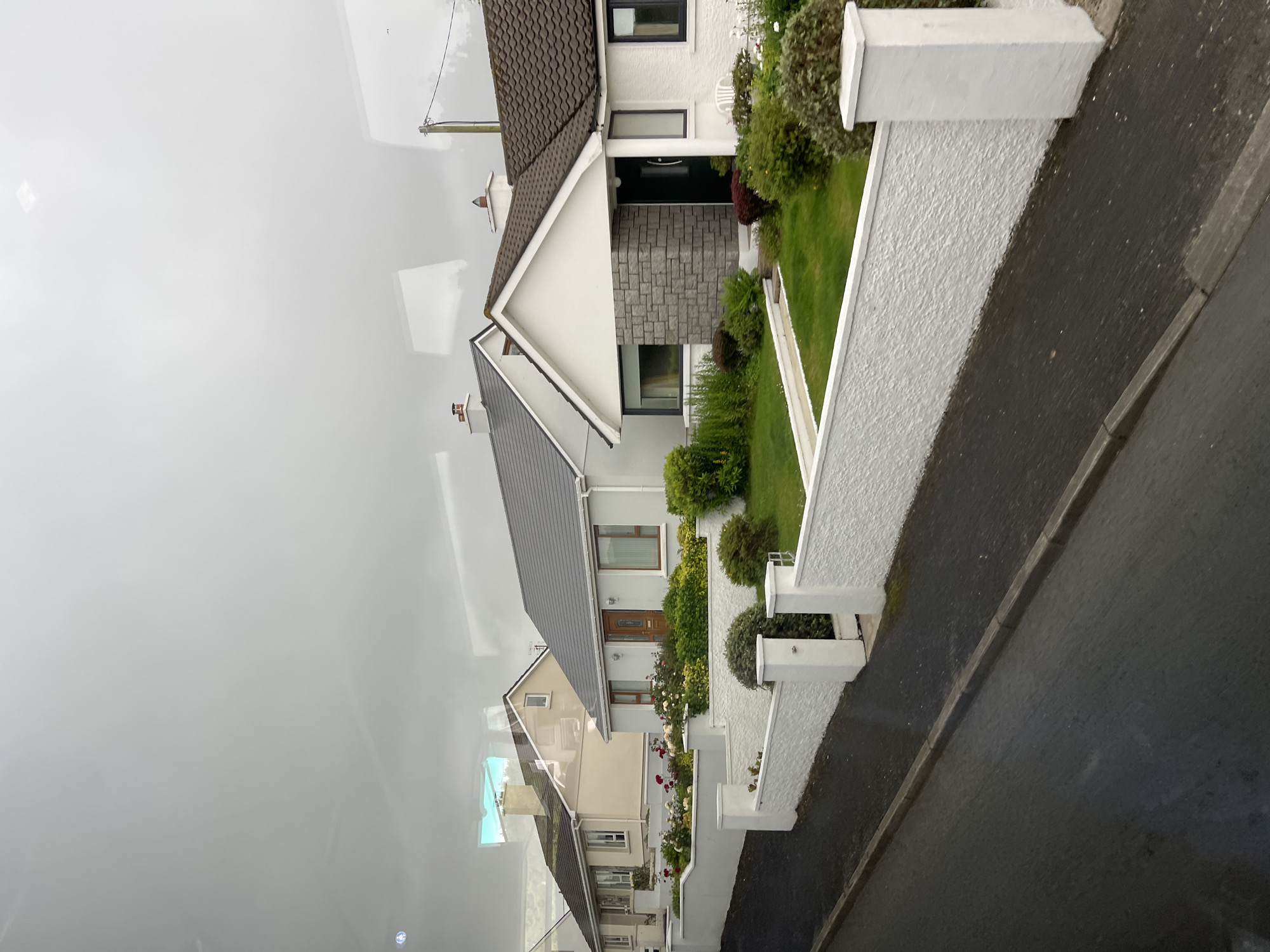 The image shows a row of detached houses on a street in Falcarragh, Ireland. The houses are of a similar style, featuring pitched roofs, white or light-colored walls, and gardens. The street in front of the houses is paved with asphalt and has a concrete curb. A white textured wall with square pillars borders the gardens of the houses. The sky is overcast, and there are reflections on what appears to be a window, possibly from a vehicle. The houses are well-maintained with manicured lawns and flowerbeds. One house has a prominent stone facade around its entrance. Telephone poles and wires are visible in the background.