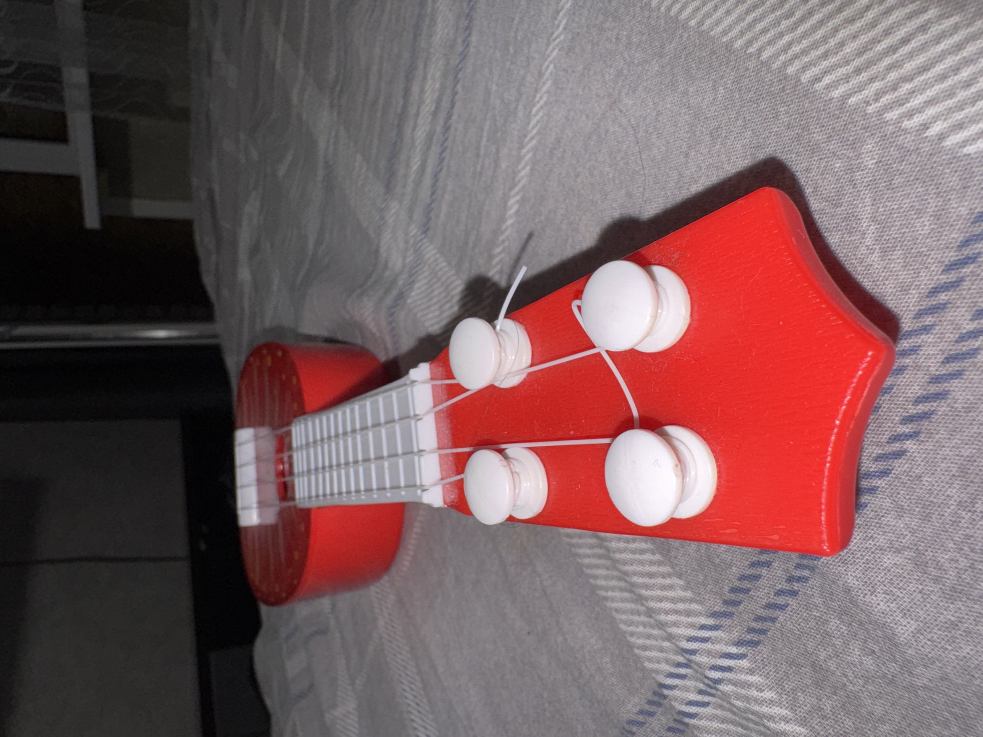 A close-up, low-angle shot shows the headstock and tuning pegs of a bright red toy ukulele. The ukulele is positioned on a grey and white striped fabric surface, possibly a bedspread or tablecloth. Four white tuning pegs are visible, each with a string wrapped around it. The frets of the ukulele are also visible, appearing white and thin. The background is dark and out of focus, with hints of furniture and a window with curtains. There are no people present in the image. The scene appears to be indoors.