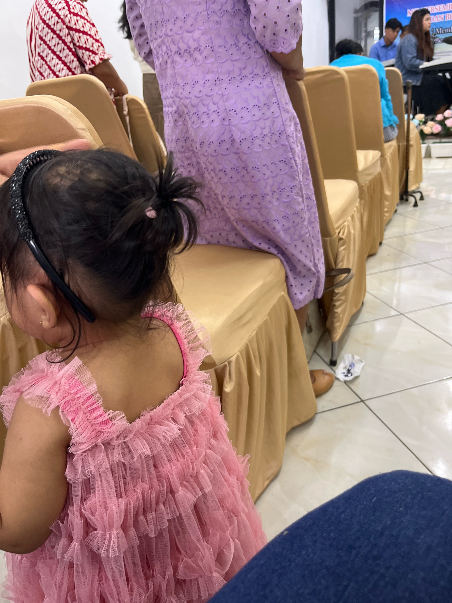 The image depicts an indoor scene with several people seated on chairs covered with gold-colored fabric. In the foreground, a young child wearing a pink frilly dress with ruffles on the sleeves and bodice is partially visible, facing away from the camera. The child has dark hair pulled into a bun and is wearing a black headband. Behind the child, several rows of chairs are visible, some occupied by individuals. One person in the middle ground is wearing a light purple dress with an intricate, textured pattern. Another person is wearing a red and white striped shirt. Further back, other individuals are seated, and a banner with text is visible in the upper right corner. The floor is tiled with a light-colored pattern. The overall setting suggests a gathering or event. The location is Marindal 1, Indonesia.
